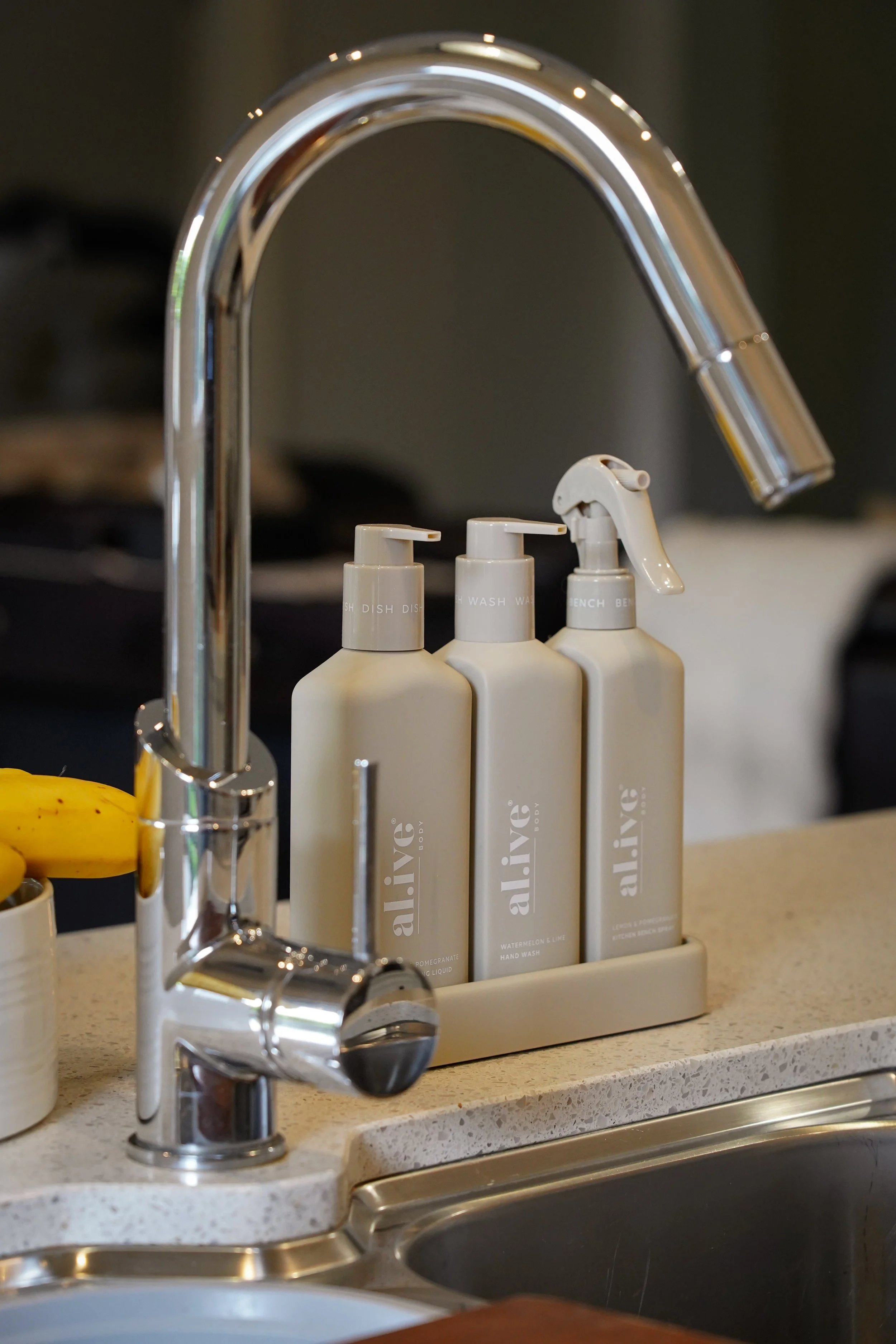 Three bottles of Alice brand soap or lotion on a beige kitchen sink counter, with a shiny chrome faucet and a small white container with bananas inside.