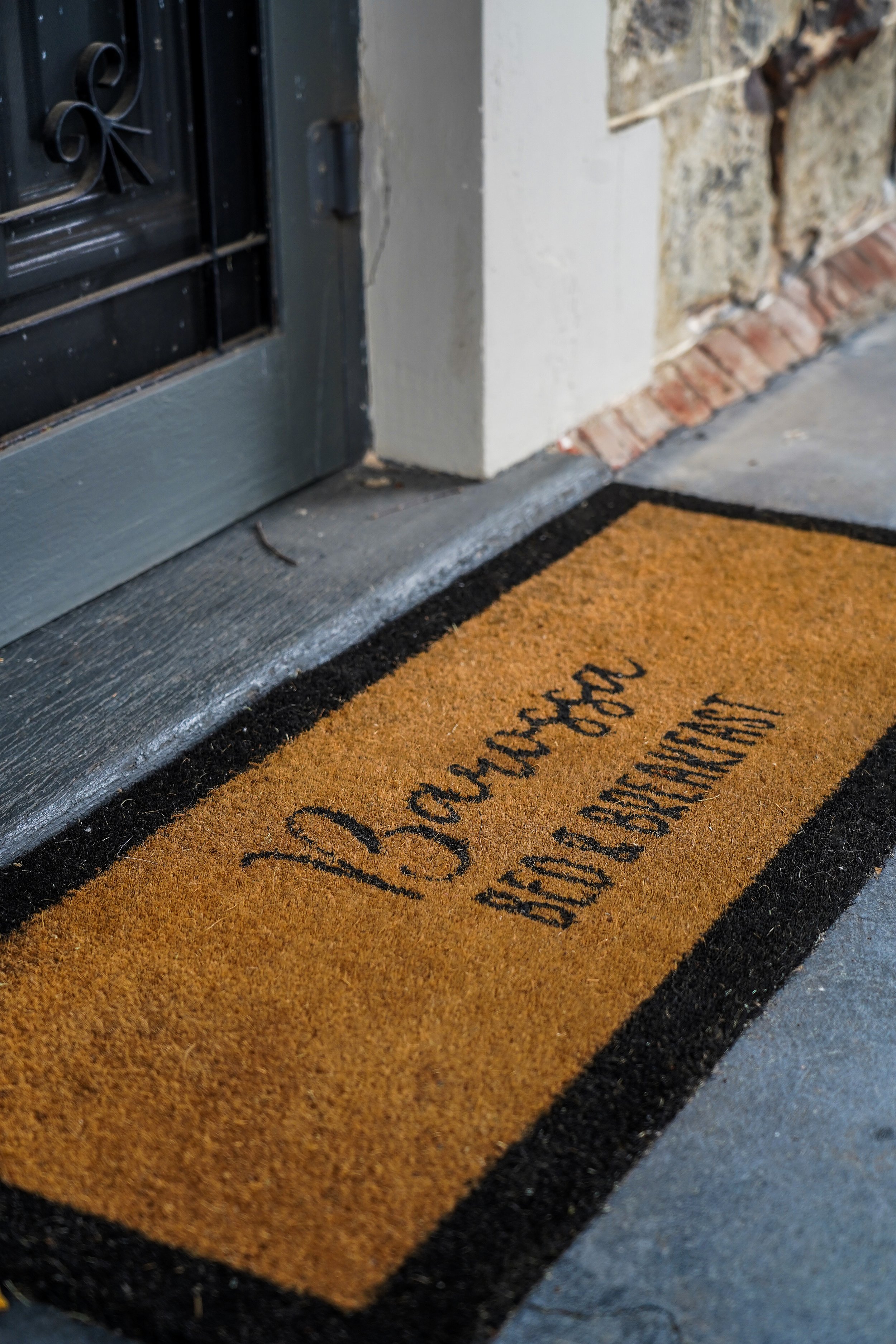 A welcome doormat with orange and black colors, reading 'Welcome' in cursive and 'La Casa de [illegible]' in uppercase, placed outside a house near a black door with a metal grille and a stone wall.