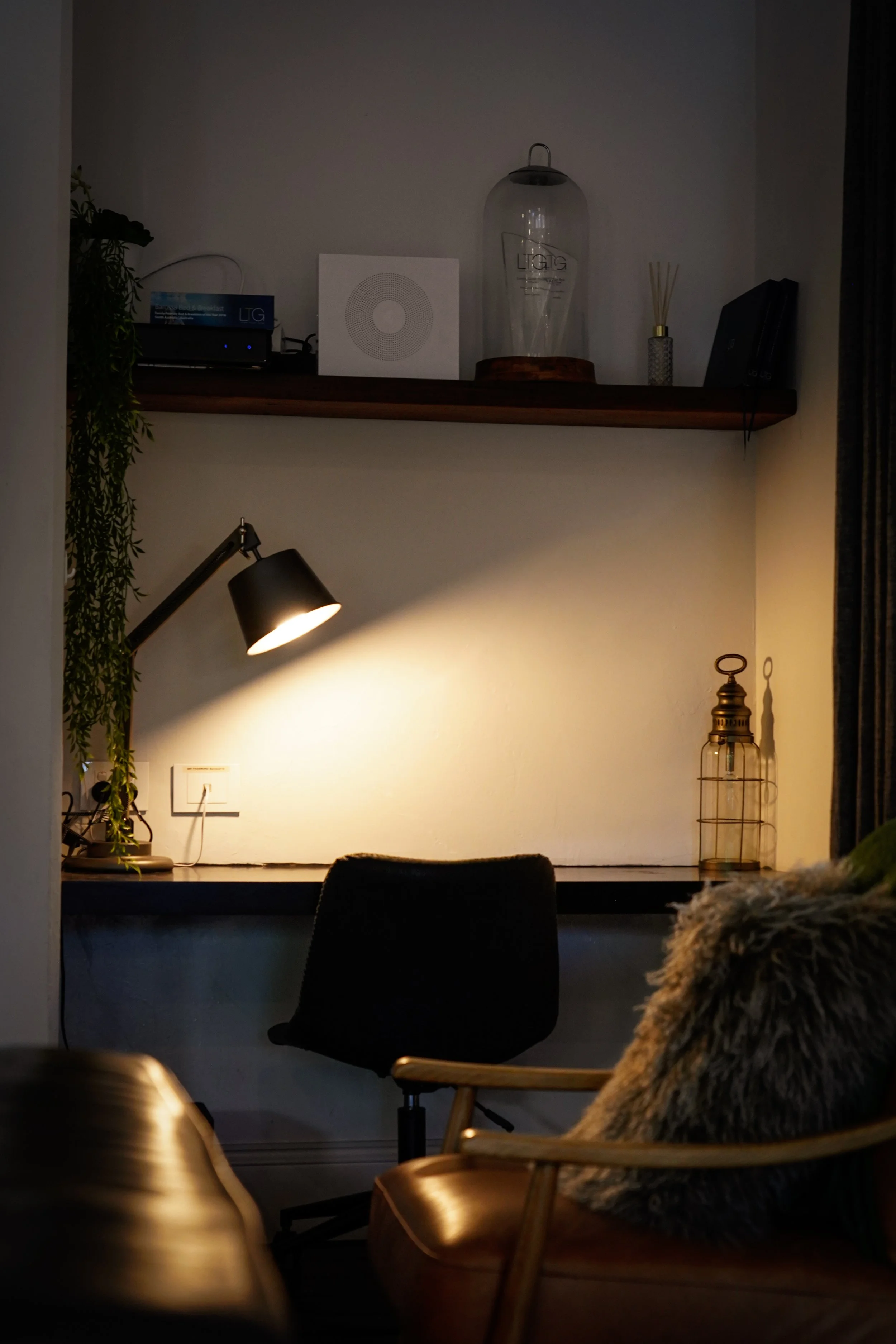 A cozy workspace with a dark desk, black office chair, and a lamp illuminating the area. Shelves above hold decorative objects, a diffuser, and a small plant. In the foreground, there is a chair with a fur pillow, and part of a leather sofa is visibl