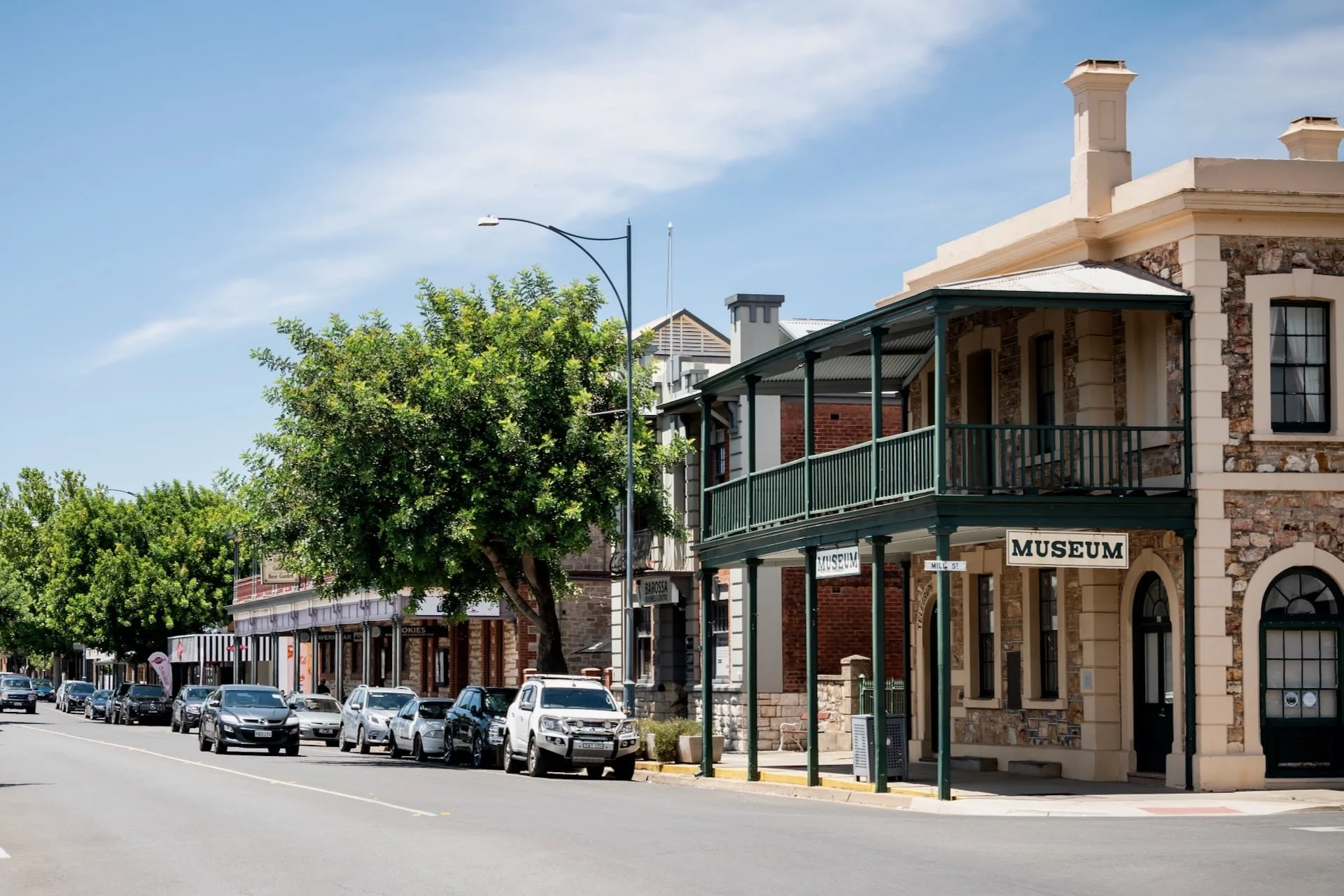 Street view of a small town with historic buildings made of brick and stone, some with signs indicating a museum. Cars are parked along the sidewalk, and there are lush green trees lining the street under a bright blue sky.