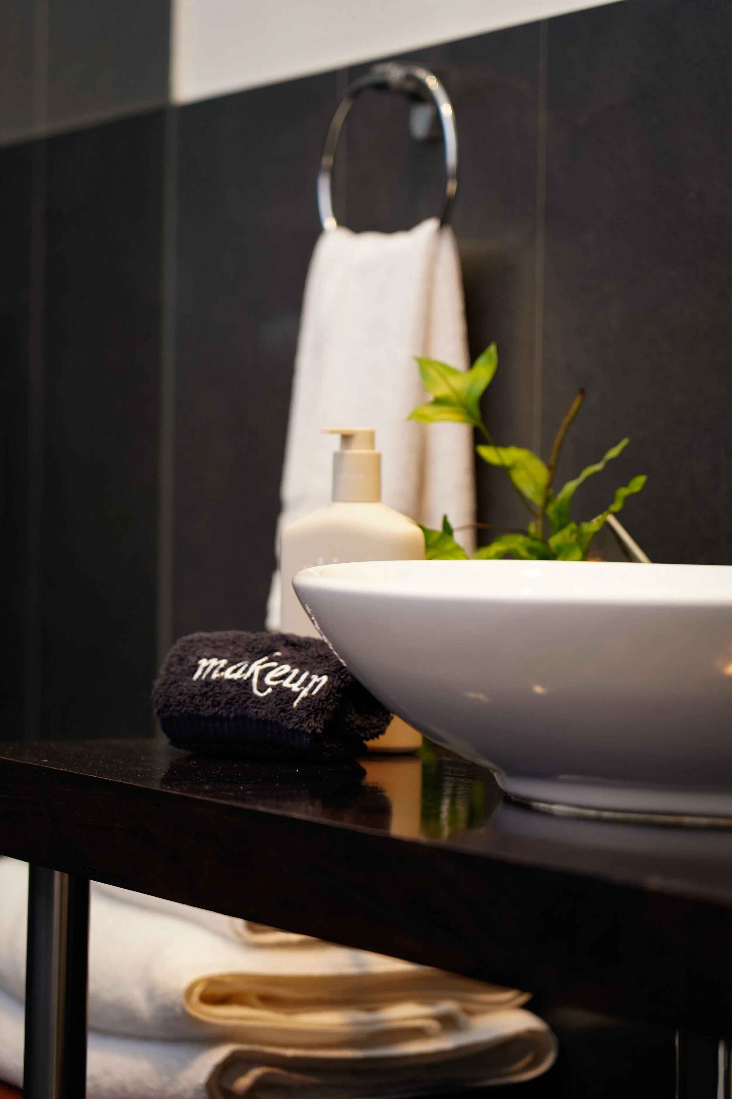 A bathroom countertop with a white vessel sink, a soap dispenser, a green plant, a white towel, a white towel ring, and a black makeup towel.