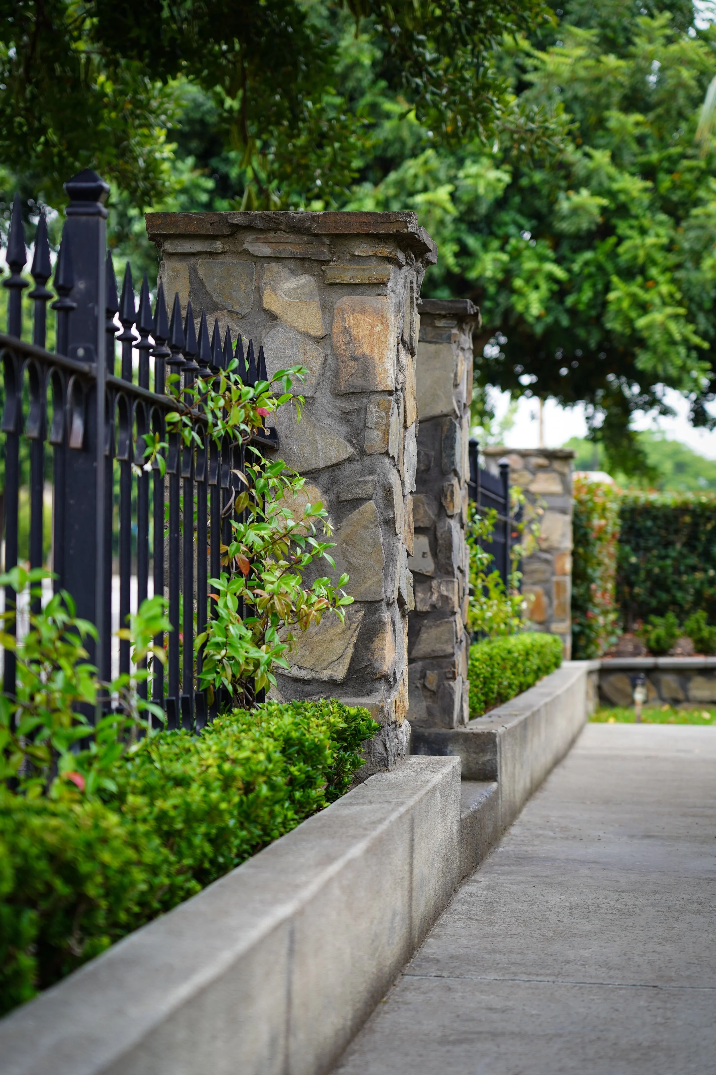 A sidewalk runs alongside a stone wall with metal fencing, surrounded by lush green bushes and trees.