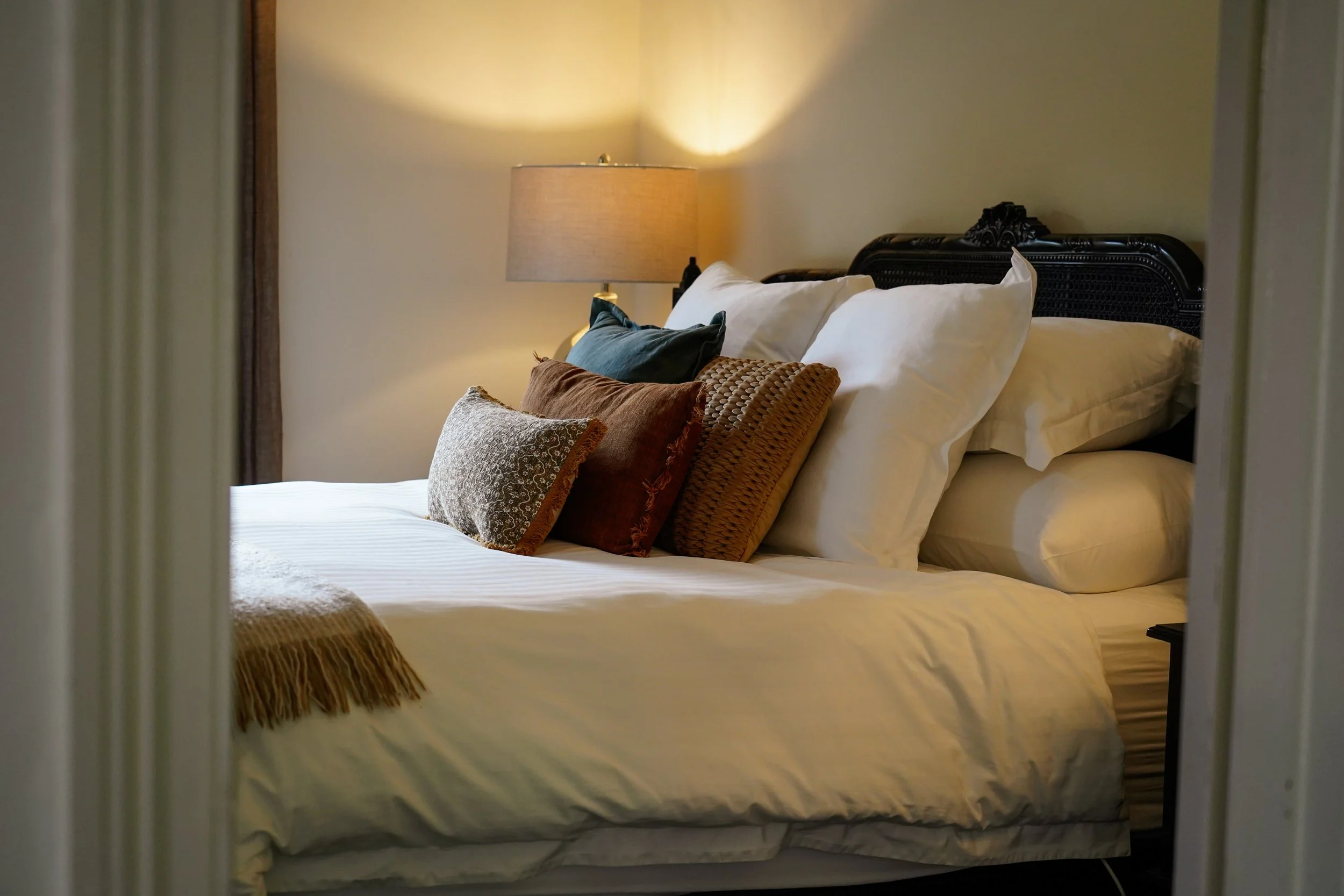 A neatly made bed with multiple pillows and a bedside lamp in a warmly lit bedroom.