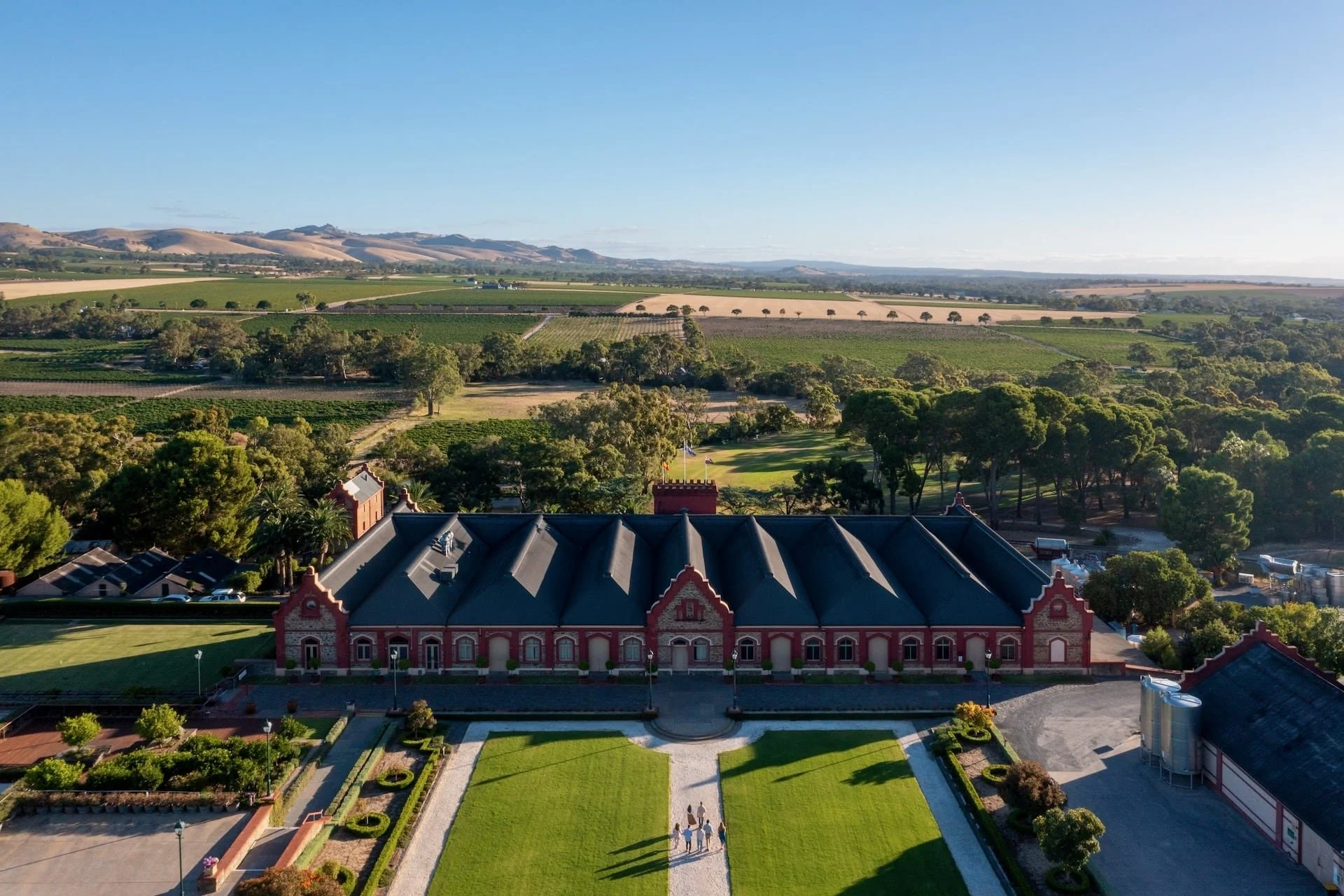 Aerial view of a historic red brick building with a dark gray roof, surrounded by well-maintained lawns and gardens, with a group of people walking toward the entrance, set against lush green fields and rolling hills in the background on a sunny day.
