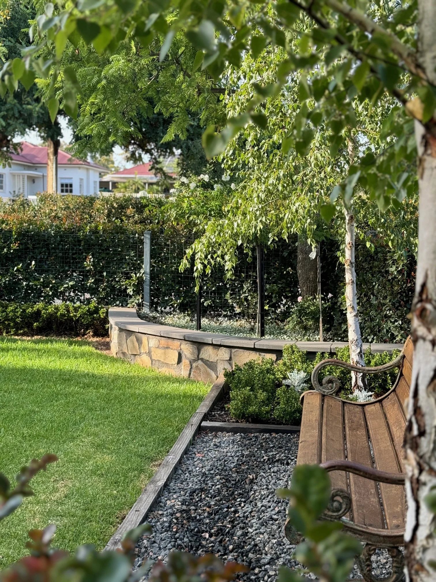 A garden scene with a wooden bench on a gravel path, surrounded by trees, bushes, and a stone wall with a metal fence in the background.