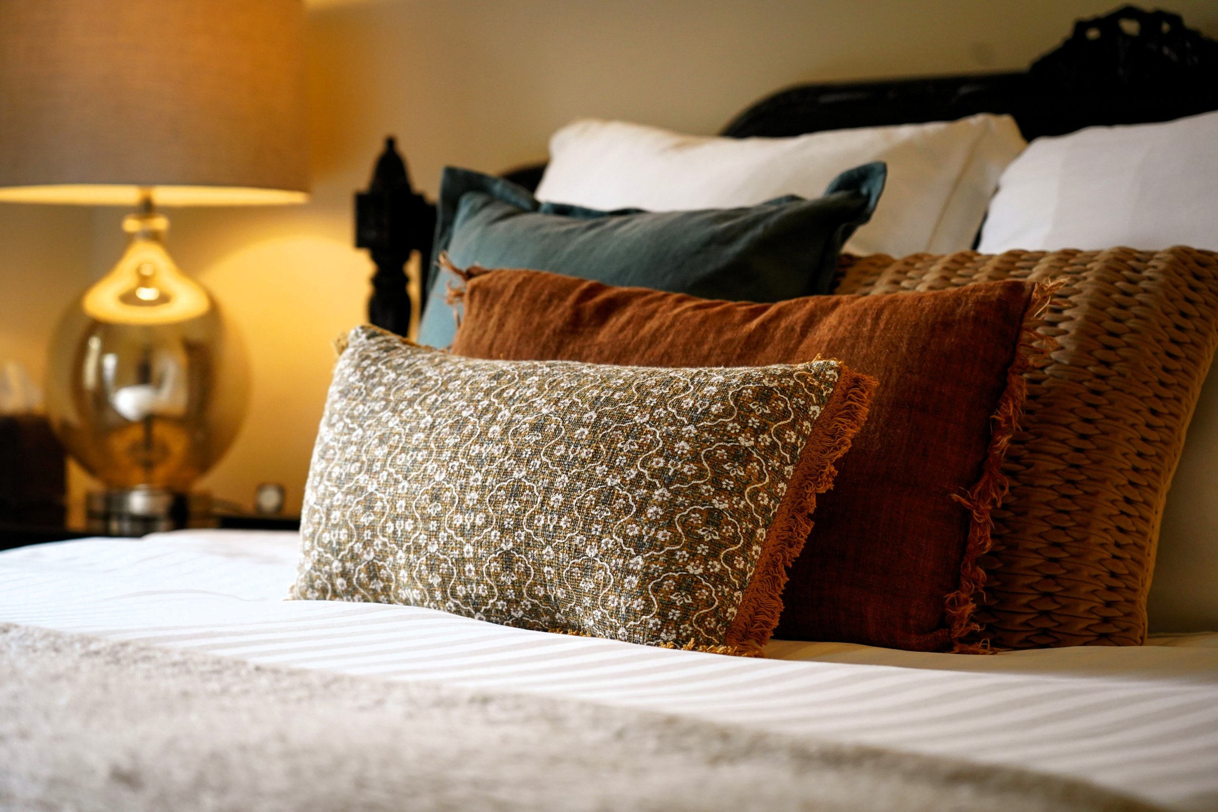 A neatly made bed with multiple decorative pillows in brown, rust, green, and white with a patterned pillow front and a white bedsheet. A side table with a gold-colored table lamp is visible in the background.