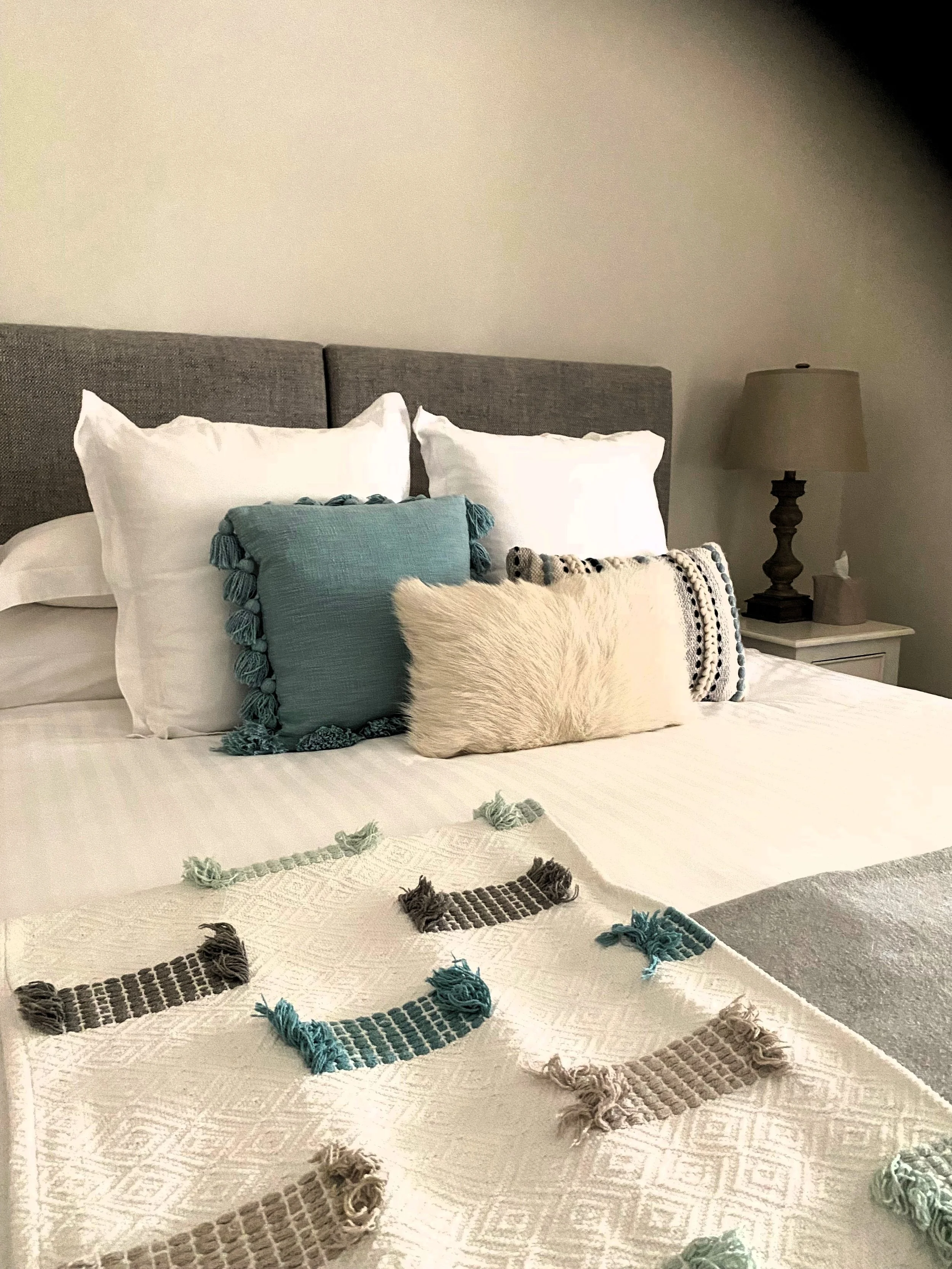 A neatly made bed with white sheets, two large white pillows, and three smaller decorative pillows with different textures and colors, including teal with tassels, white faux fur, and a patterned pillow. There is a nightstand with a beige lamp and a 