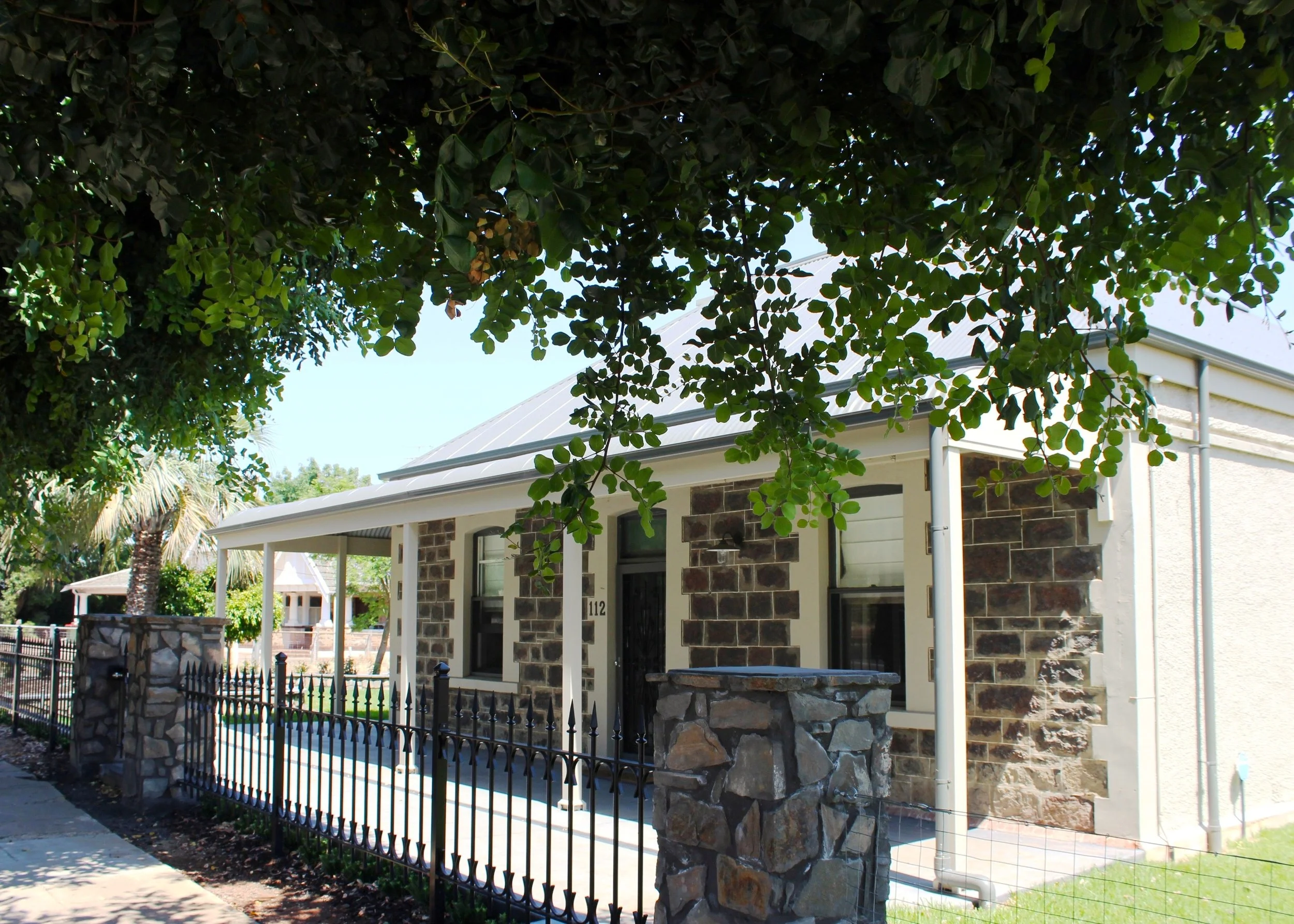 Front view of a house with stone and white siding, small porch, metal fence, and large tree with overhanging branches