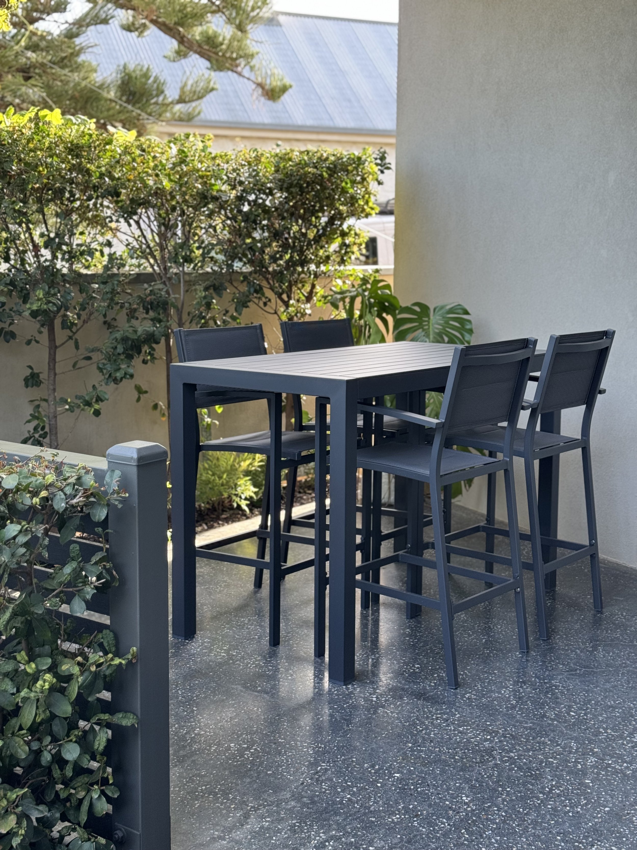 Outdoor patio with a tall black table and four matching high chairs, surrounded by greenery and a white wall.