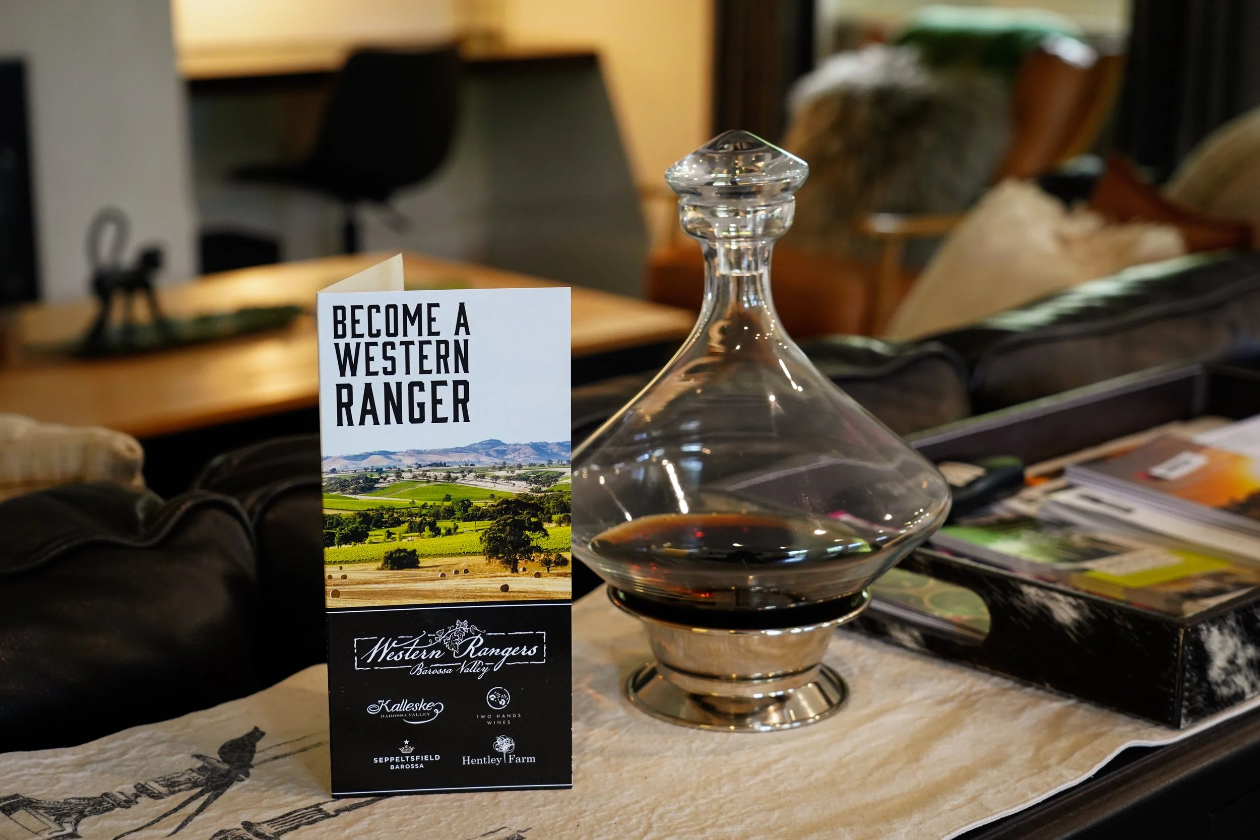 A glass decanter with dark liquid inside, placed on a table next to a promotional standee that reads 'Become a Western Ranger' with a scenic landscape, and a black box with brochures or magazines on top.