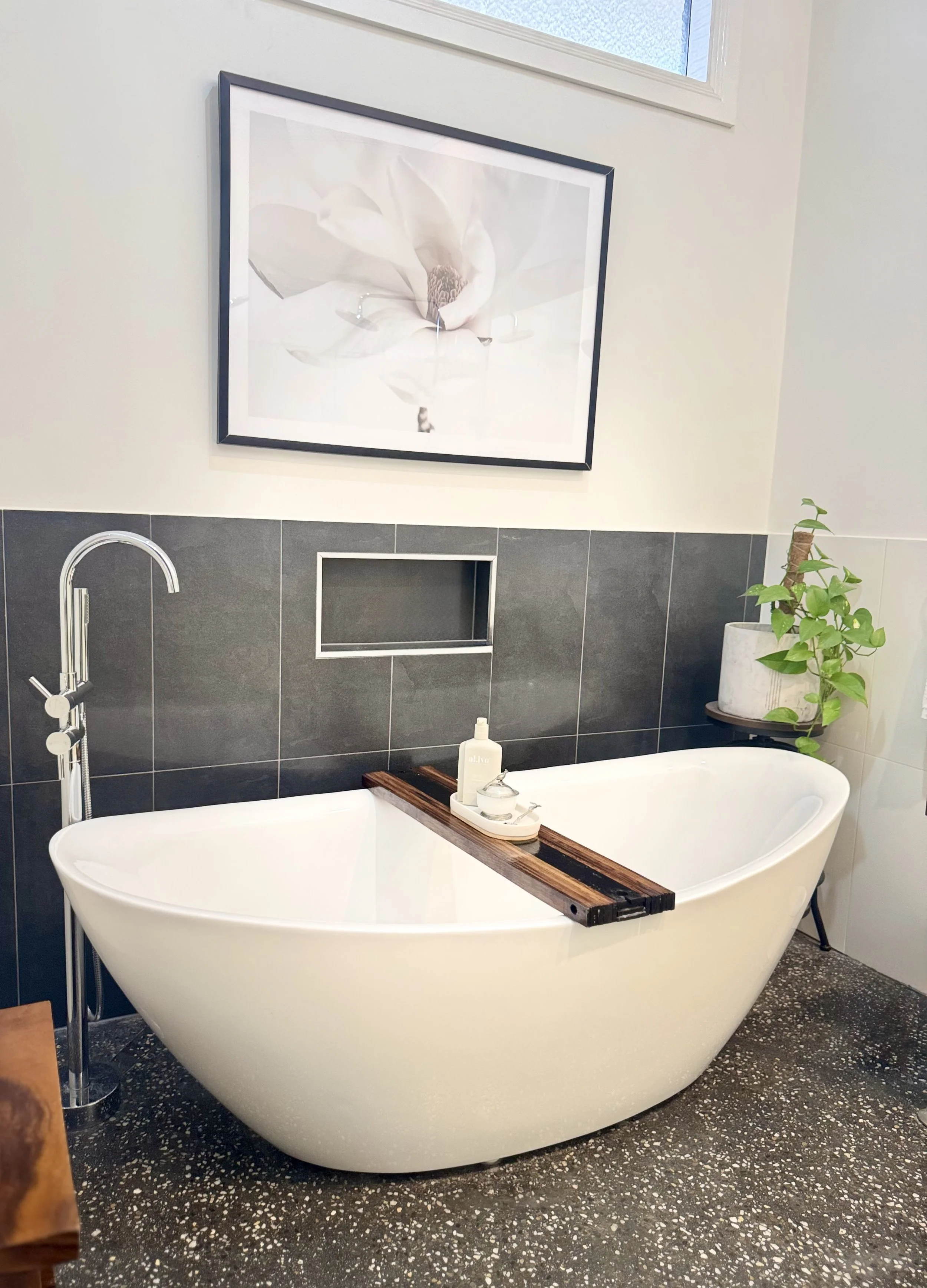 A white freestanding bathtub with a wooden bath tray, soap, and lotion bottles placed on it. A plant in a pot sits on a small stand in the corner, and a large framed flower photograph hangs above a gray tiled wall. The bathroom has a speckled black a