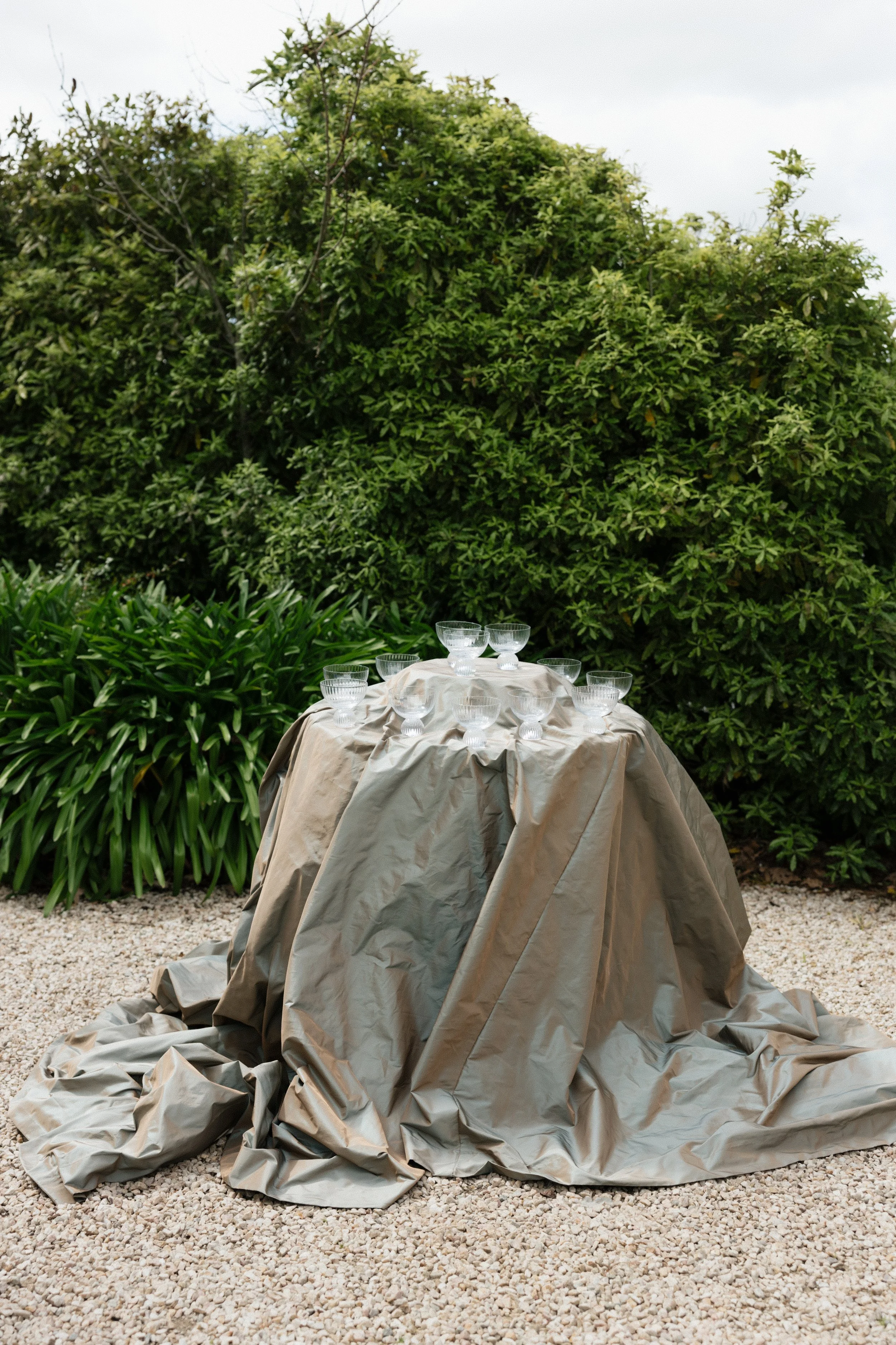 A table covered with a beige cloth, set with multiple glass bowls, outdoors on a gravel surface with lush green bushes in the background.