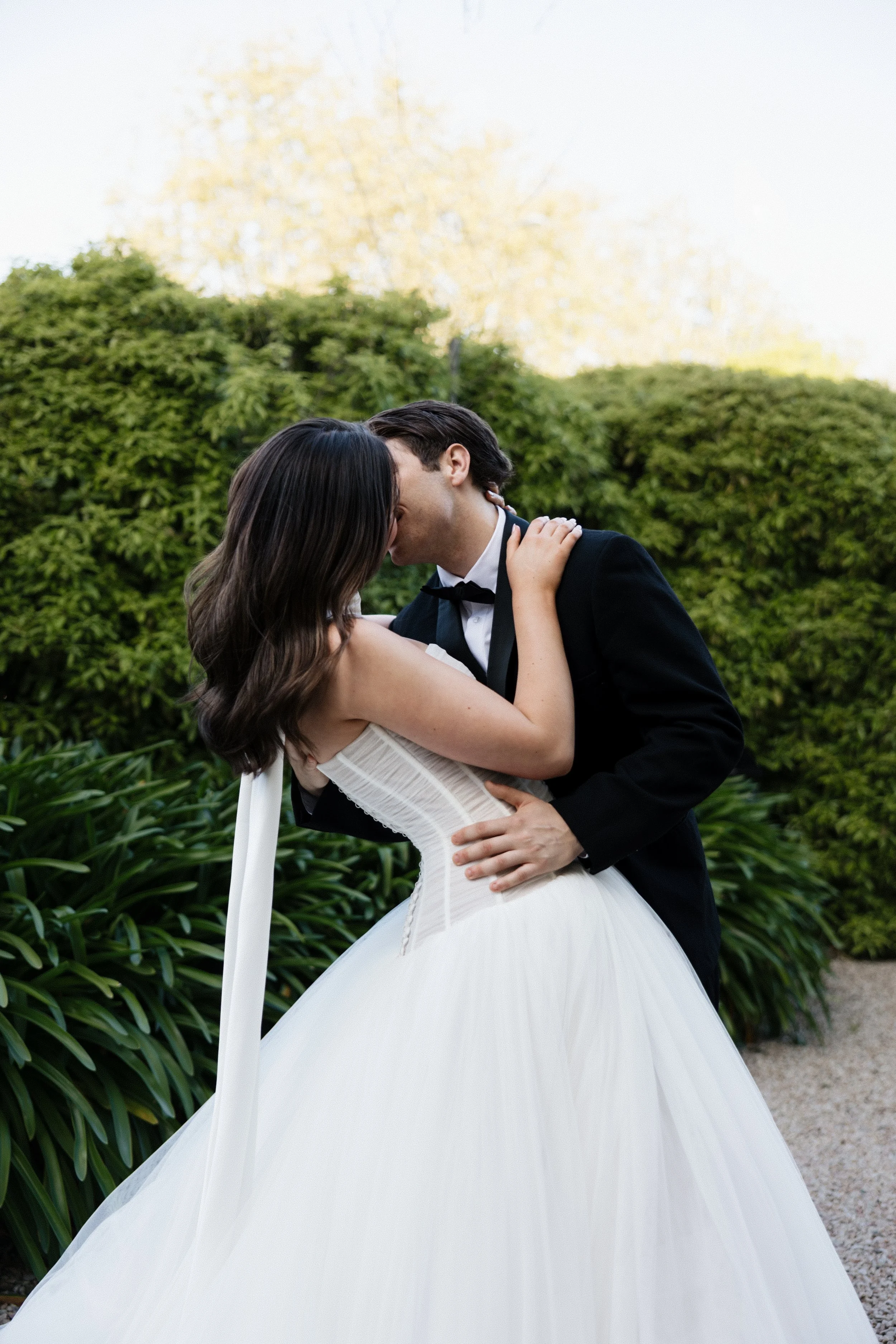 A bride and groom share a romantic kiss outdoors in a garden, with greenery and trees in the background.