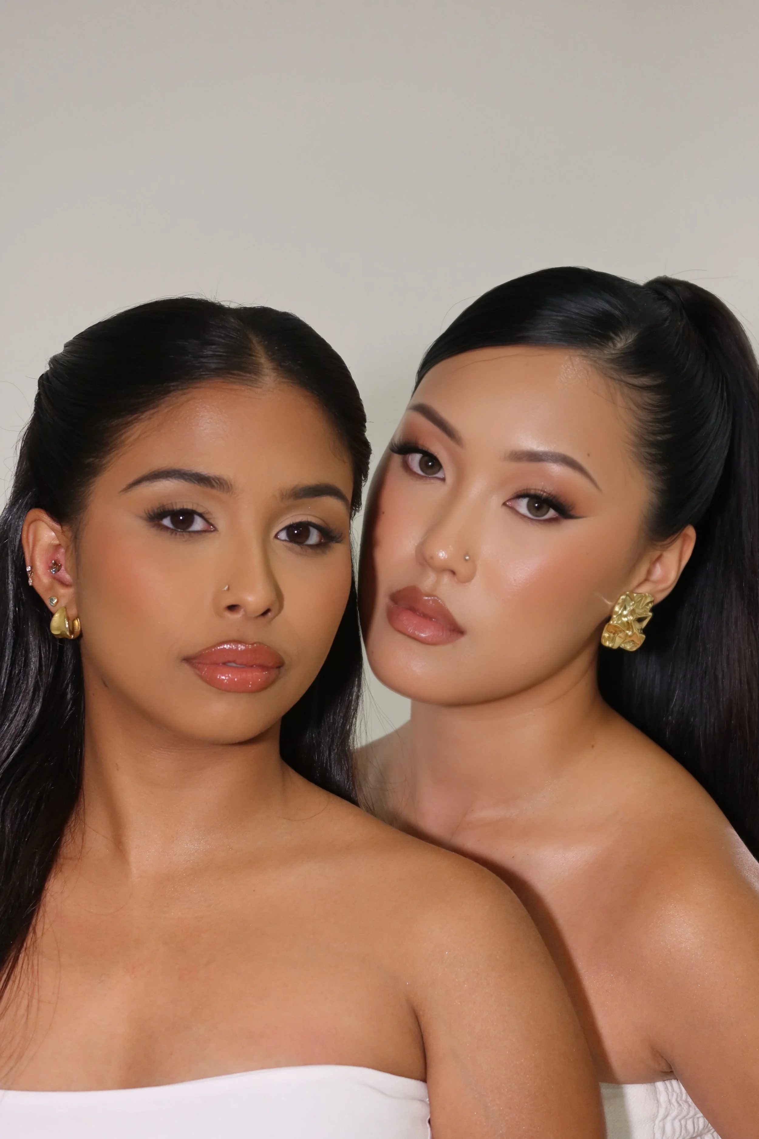Two women with dark hair and makeup posing closely together against a plain background, both wearing white and gold jewelry.