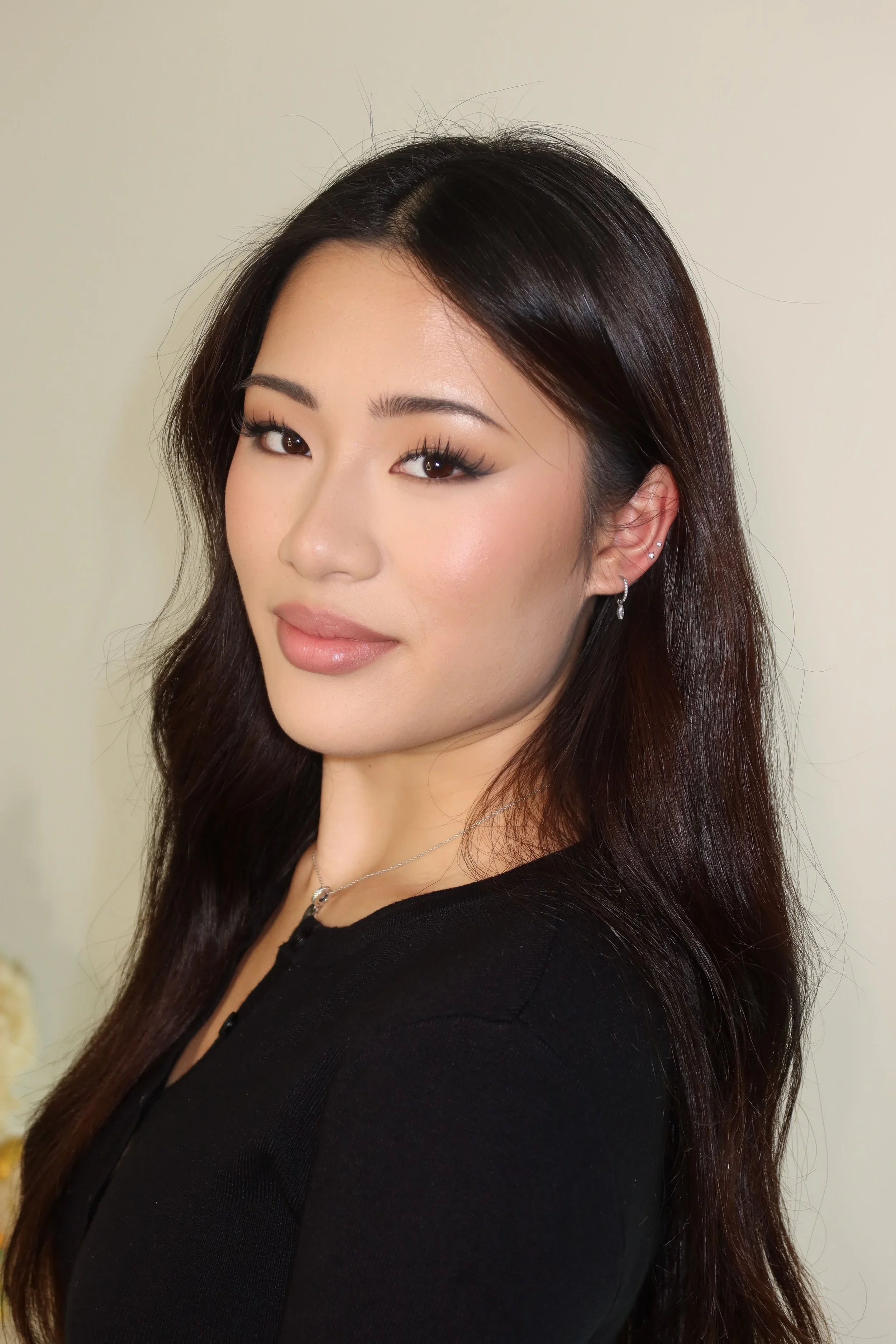 Close-up of a woman with long dark hair, wearing a black top, jewelry, and makeup, against a plain light-colored background.