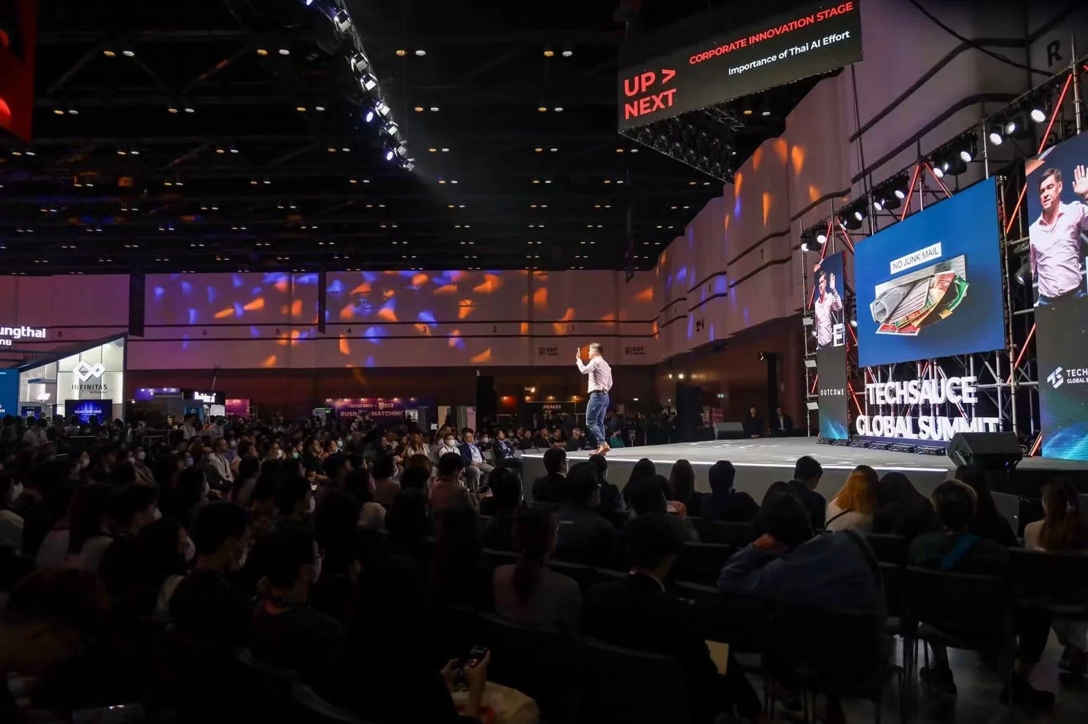 A speaker on stage at the TechSauge Global Summit giving a presentation to a large audience in a conference hall, with screens displaying visuals and the event name.