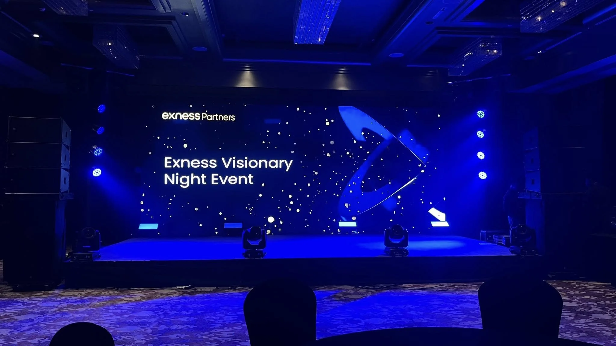 A stage with a large digital screen displaying the event name "Exness Visionary Night Event" and themed graphics, equipped with stage lights on both sides, set in a decorated conference hall.