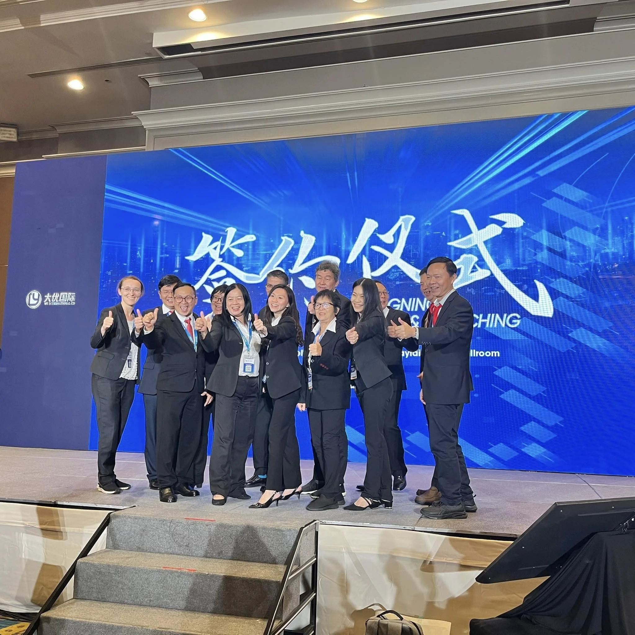 A group of eleven professionals, mostly women, dressed in formal business attire, standing on a stage in front of a large blue digital screen with Chinese characters, giving thumbs-up gestures and smiling.