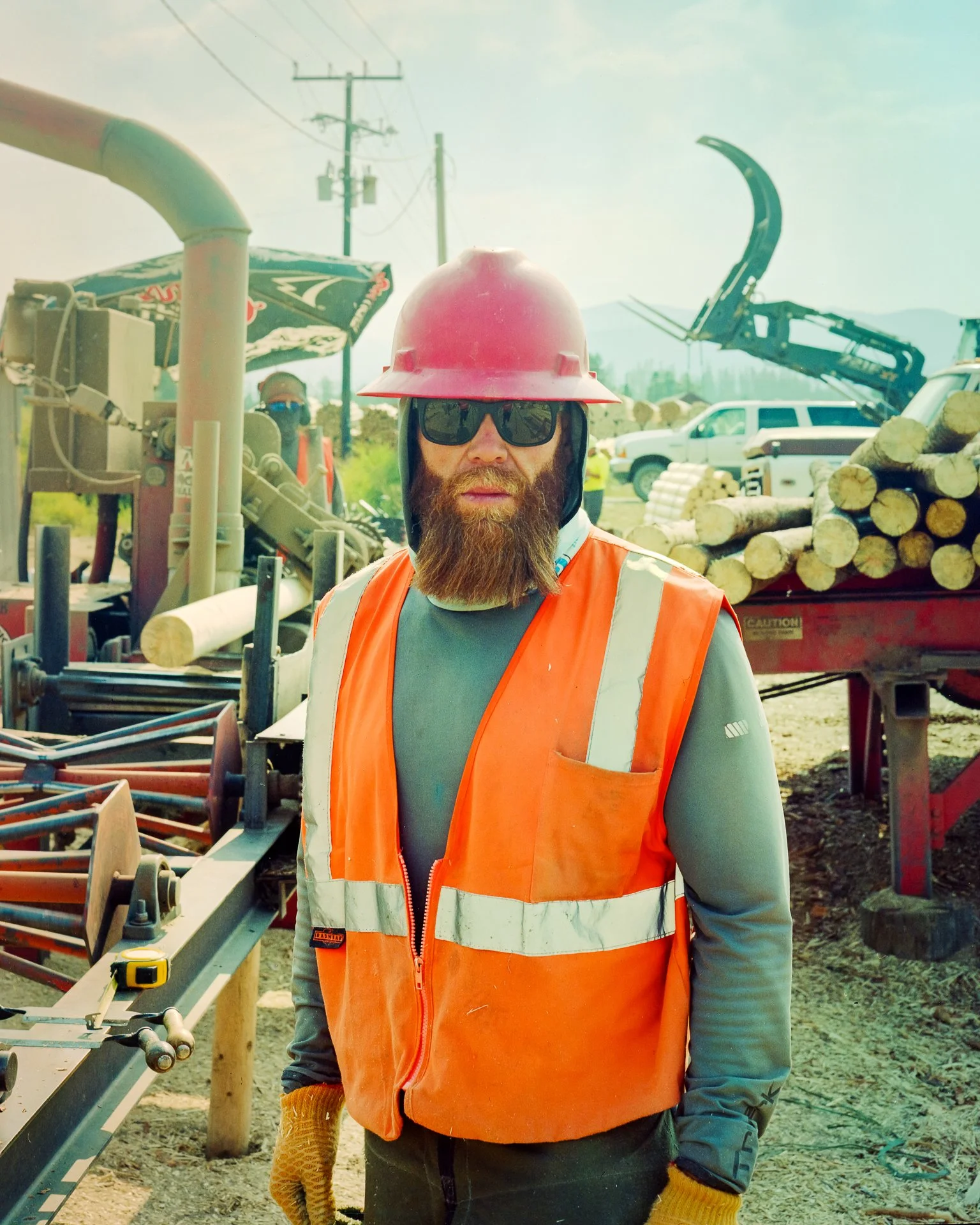 Construction worker wearing safety helmet, sunglasses, orange safety vest, and gloves at a lumber processing site with logs, machinery, and vehicles in the background.