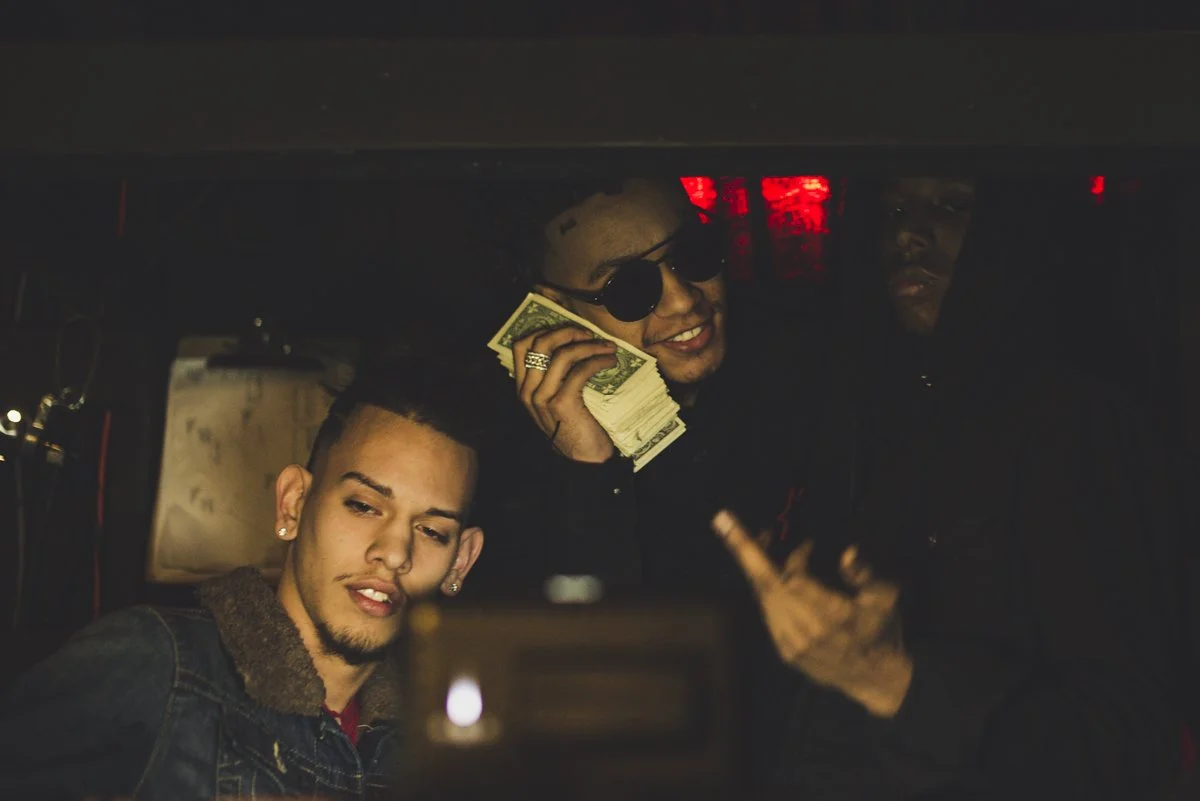 Three young men taking a selfie inside a club or bar, one holding a stack of money, wearing sunglasses, and smiling, while the other two look at the camera.