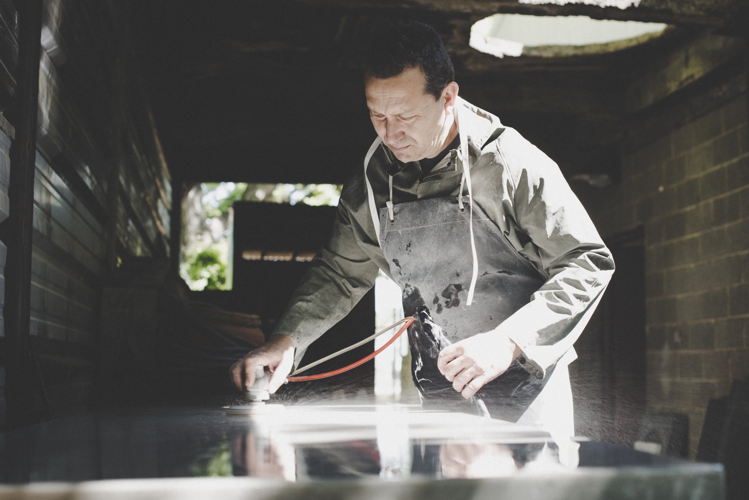 A man in a gray apron, working on a dark surface, appears to be polishing or grinding with a tool, in a workshop or garage setting.