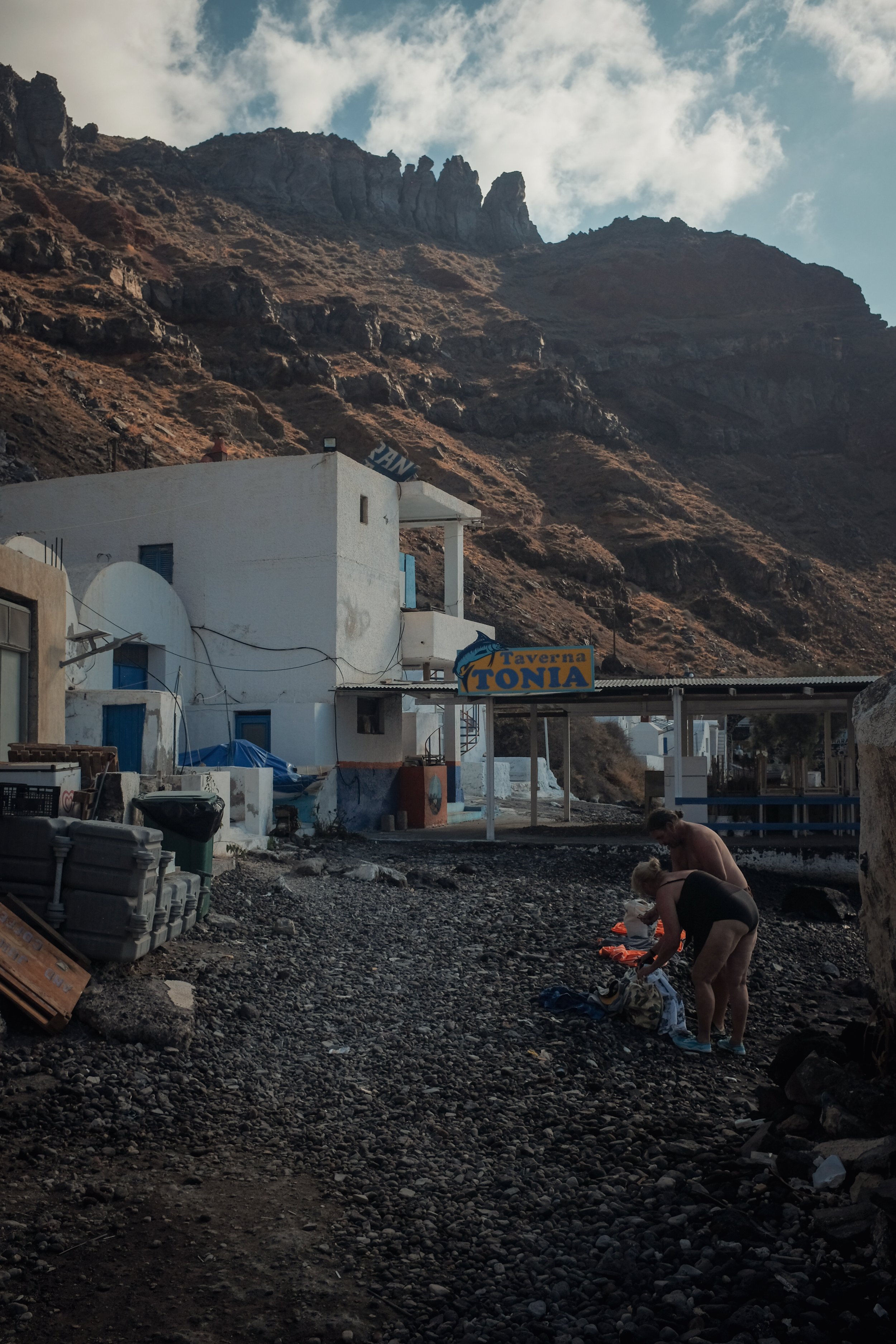 Two women, one in black shorts and the other in a black tank top, bend down on a rocky ground, sorting through clothes outdoors. Behind them, there are white buildings with blue doors and windows and a sign that reads 'Taverna Tonia.' Mountainous ter
