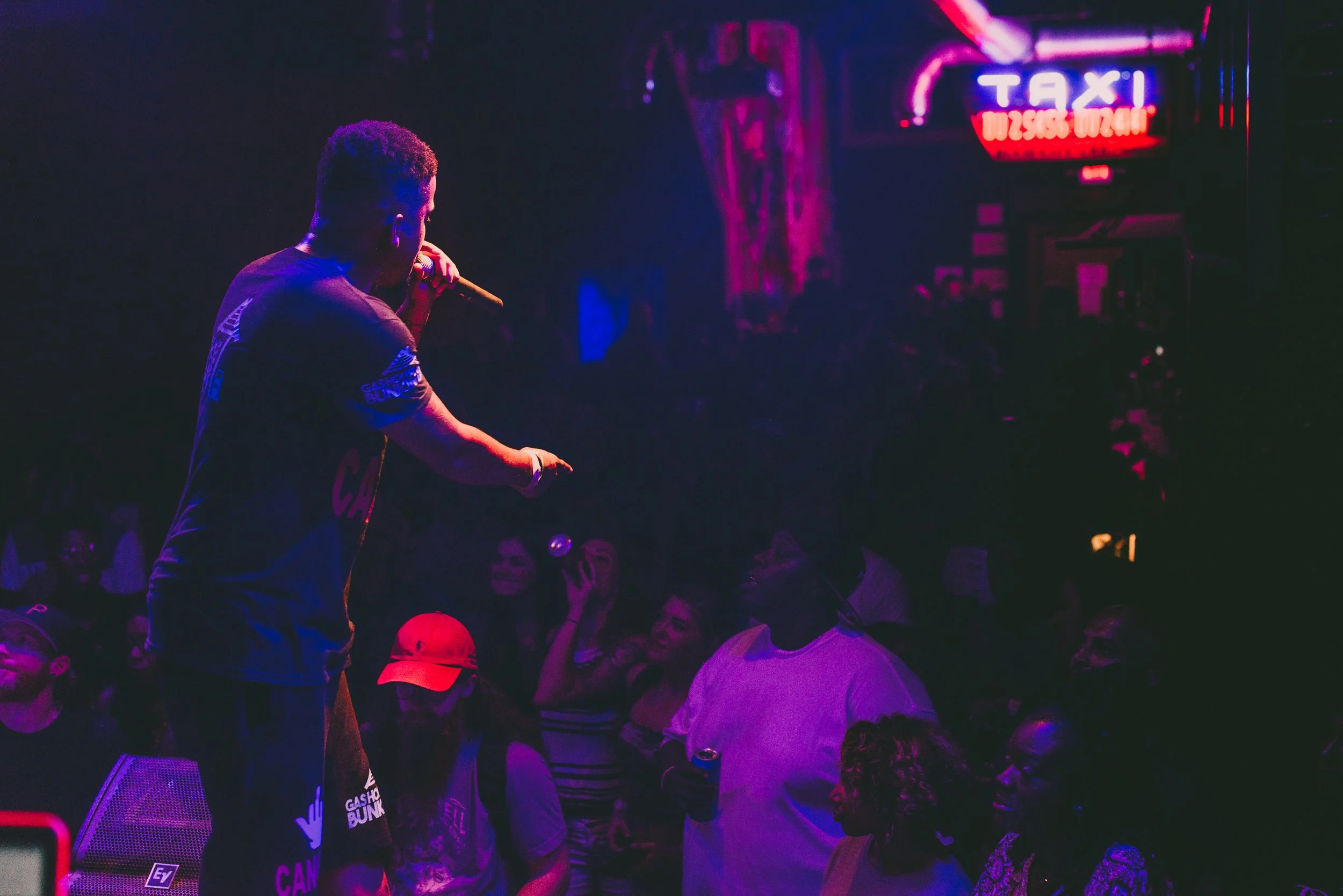A male performer or speaker holding a microphone on stage at a nightclub or concert venue, with a neon-lit 'TAXI' sign in the background. A crowd of people watching him is visible in the dark.