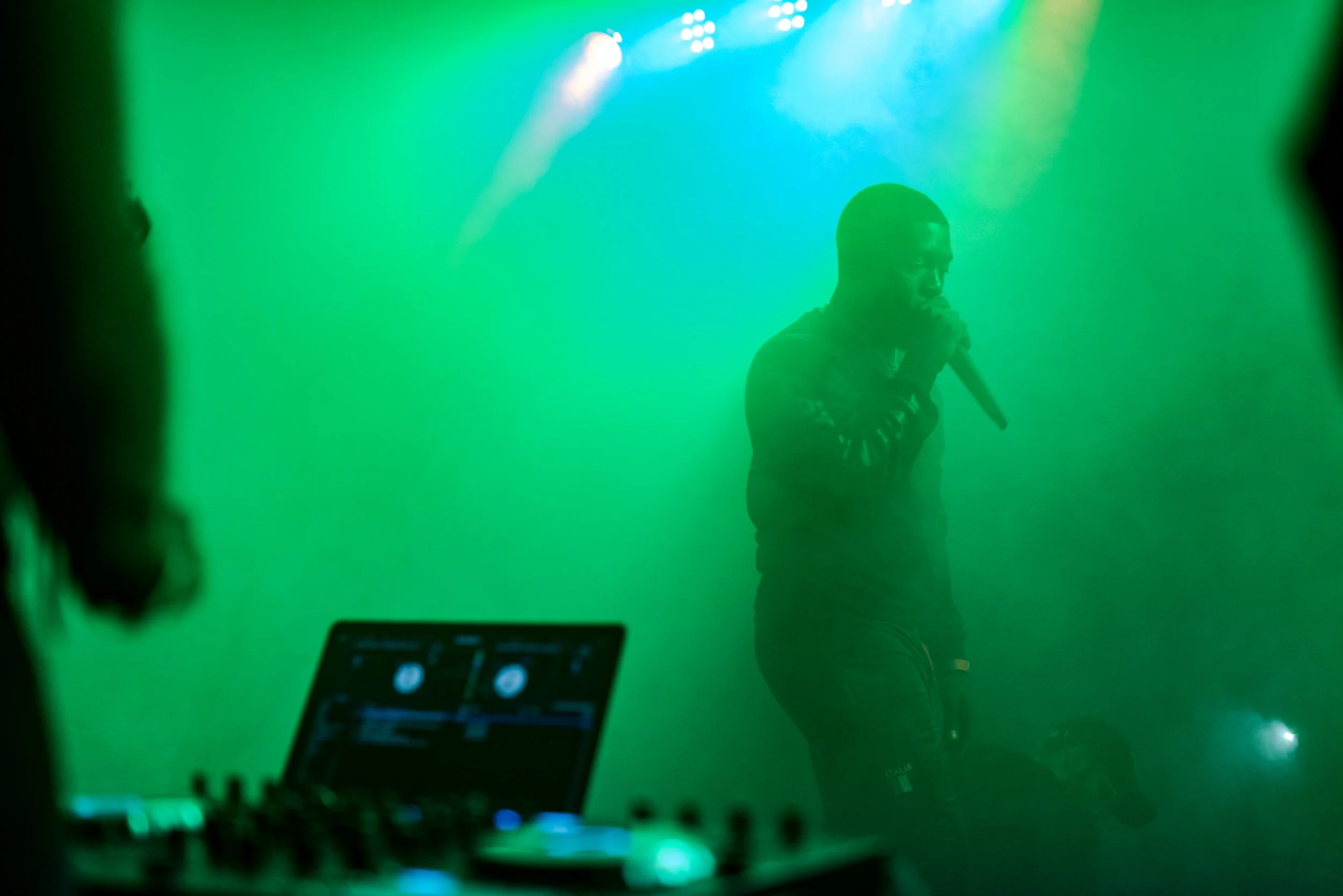 A performer holding a microphone on a green-lit stage, with a DJ console visible in the foreground.
