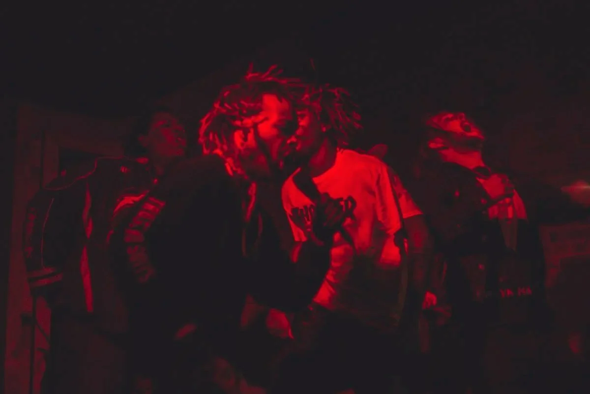Group of young people with dreadlocks and casual clothing in a dark setting illuminated by red lighting.