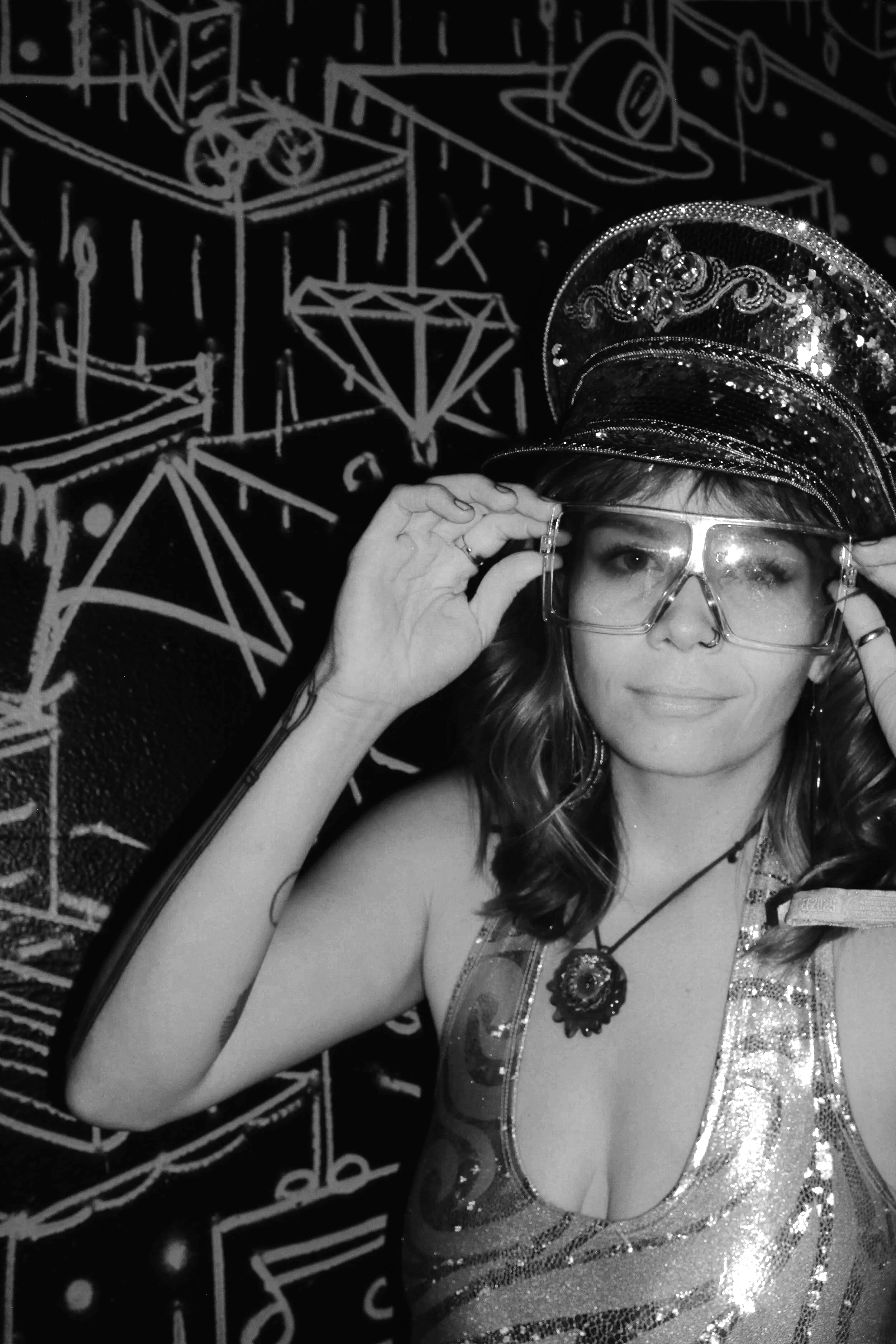 A woman wearing a shiny, sparkly top, large glasses, and a decorated cap, posing in front of a chalkboard with musical notes and geometric designs.