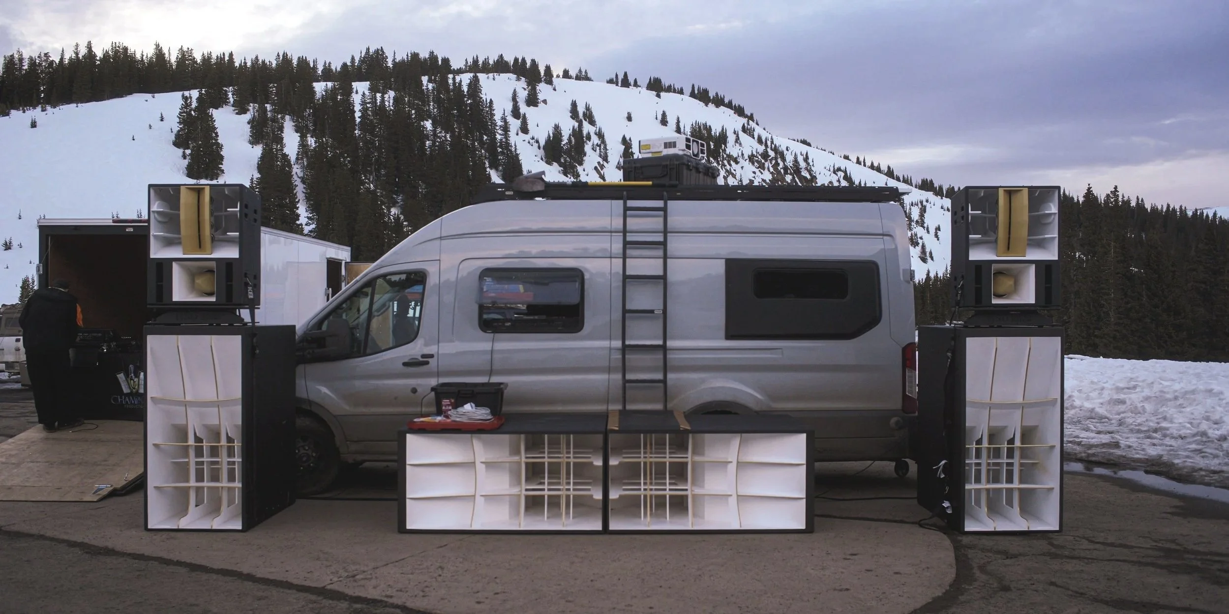 A mobile sound system setup with large speakers on either side of a gray van, which has sound equipment and a ladder on its side, set against a snowy mountain landscape.