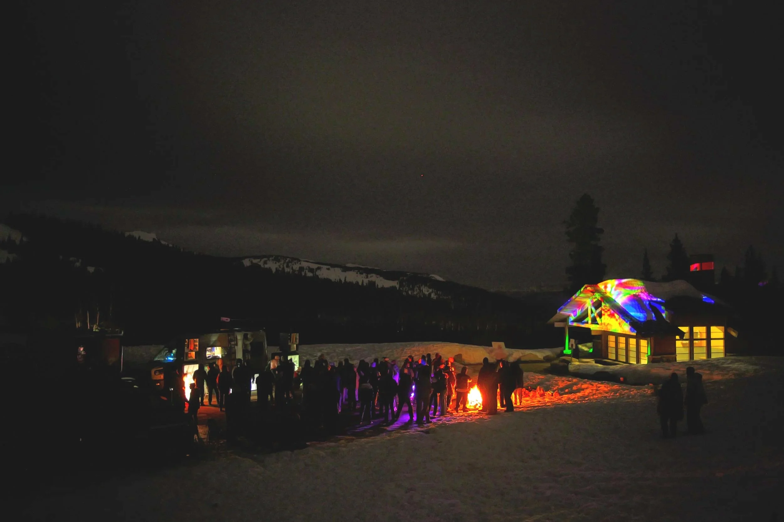 Nighttime gathering outside in snow with a campfire and colorful lights projected on a house, mountains in the background.