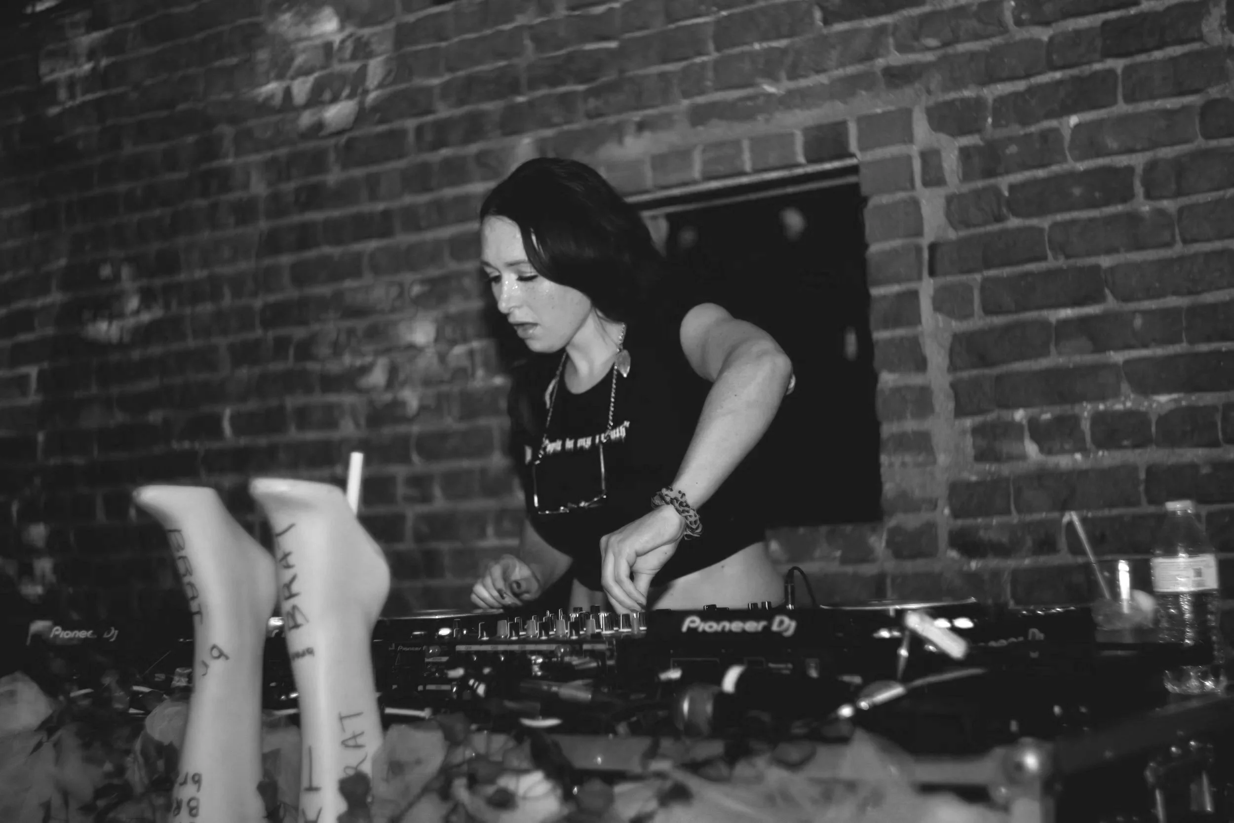 A woman DJing in a dimly lit venue with brick walls, using Pioneer DJ equipment surrounded by plants and drinks.