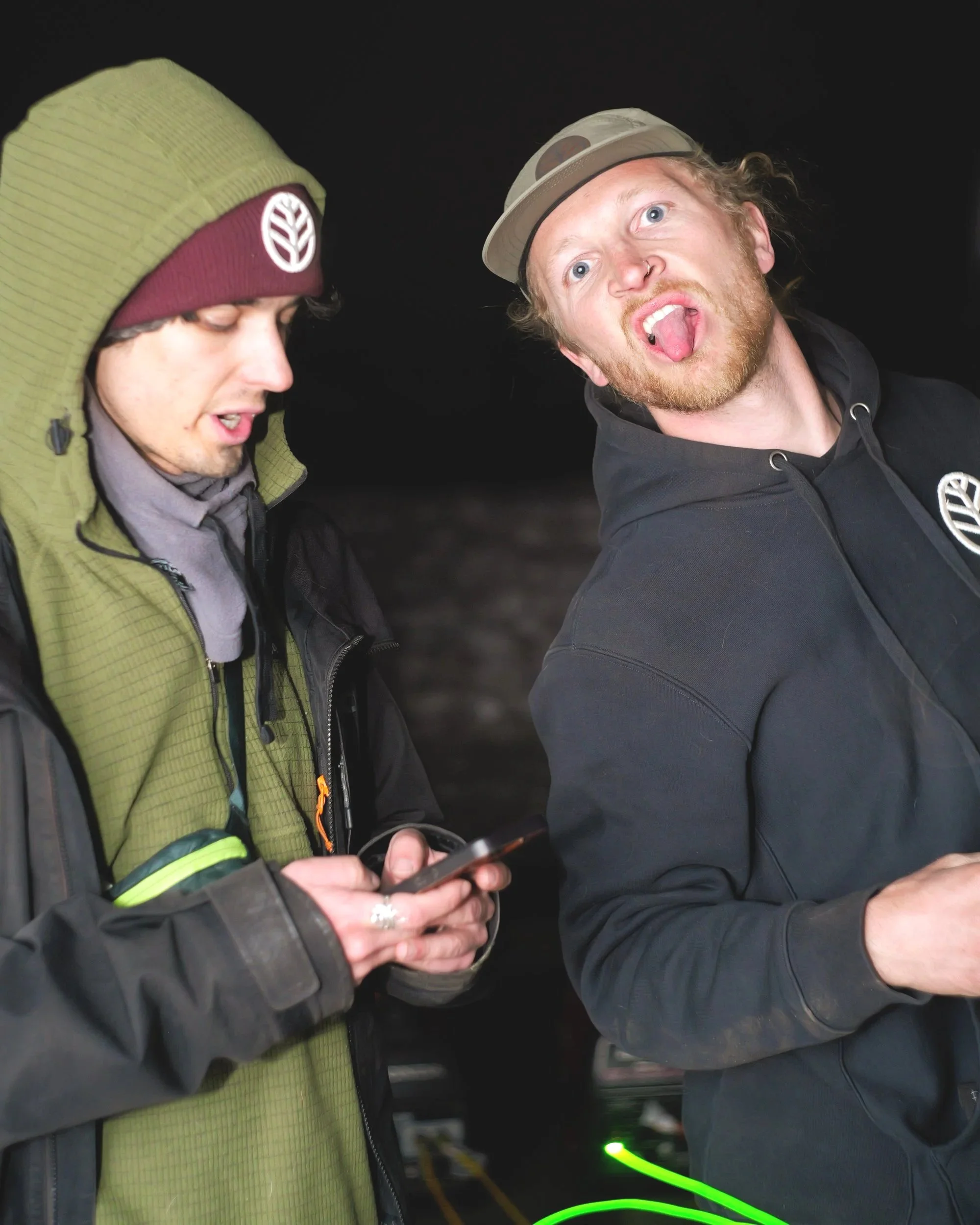 Two young men with casual clothing and beanies, one looking at a phone and the other sticking out his tongue, standing outdoors at night.