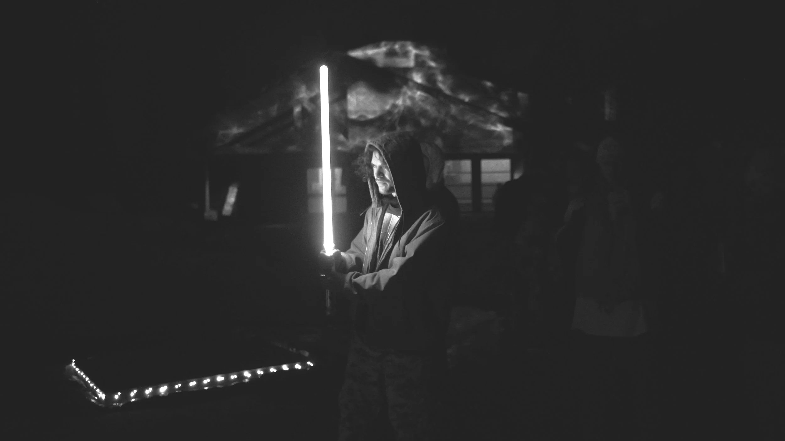 A person holding a glowing light saber in a dark outdoor setting, with a mountain in the background and faintly visible people nearby.