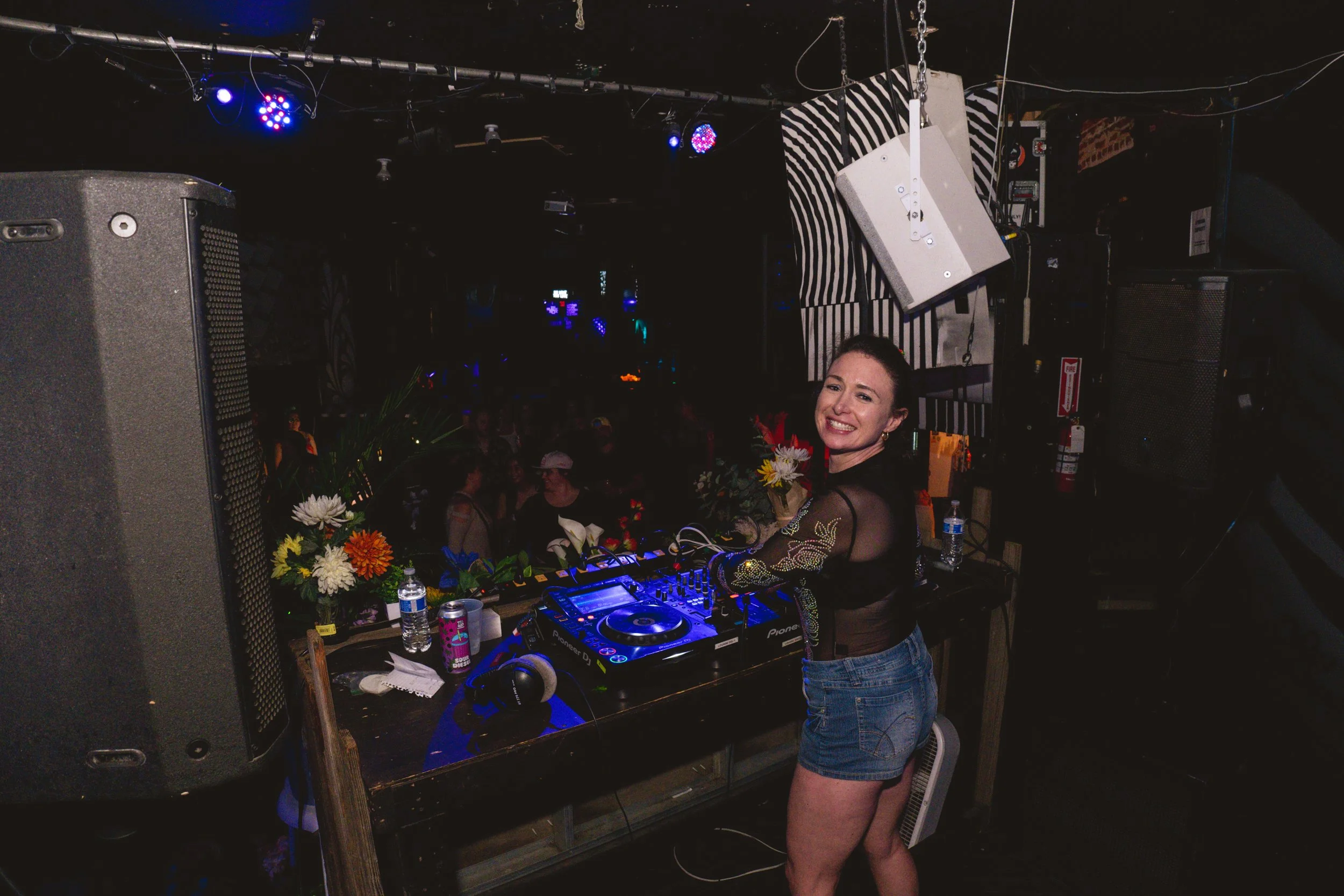 A female DJ smiling while standing at a DJ booth with speakers, flowers, water bottles, and DJ equipment in a dimly lit nightclub with people dancing in the background.
