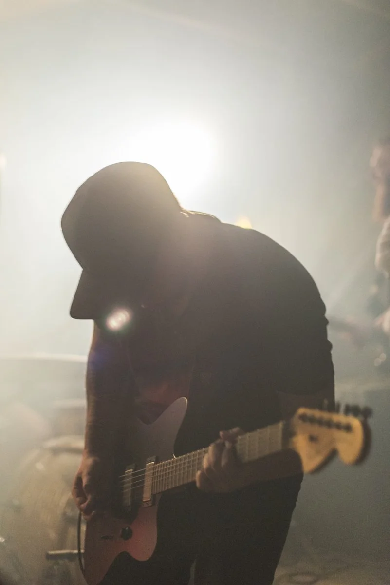 A silhouette of a person with long hair playing an electric guitar on stage with backlighting, creating a bright halo effect around their head.
