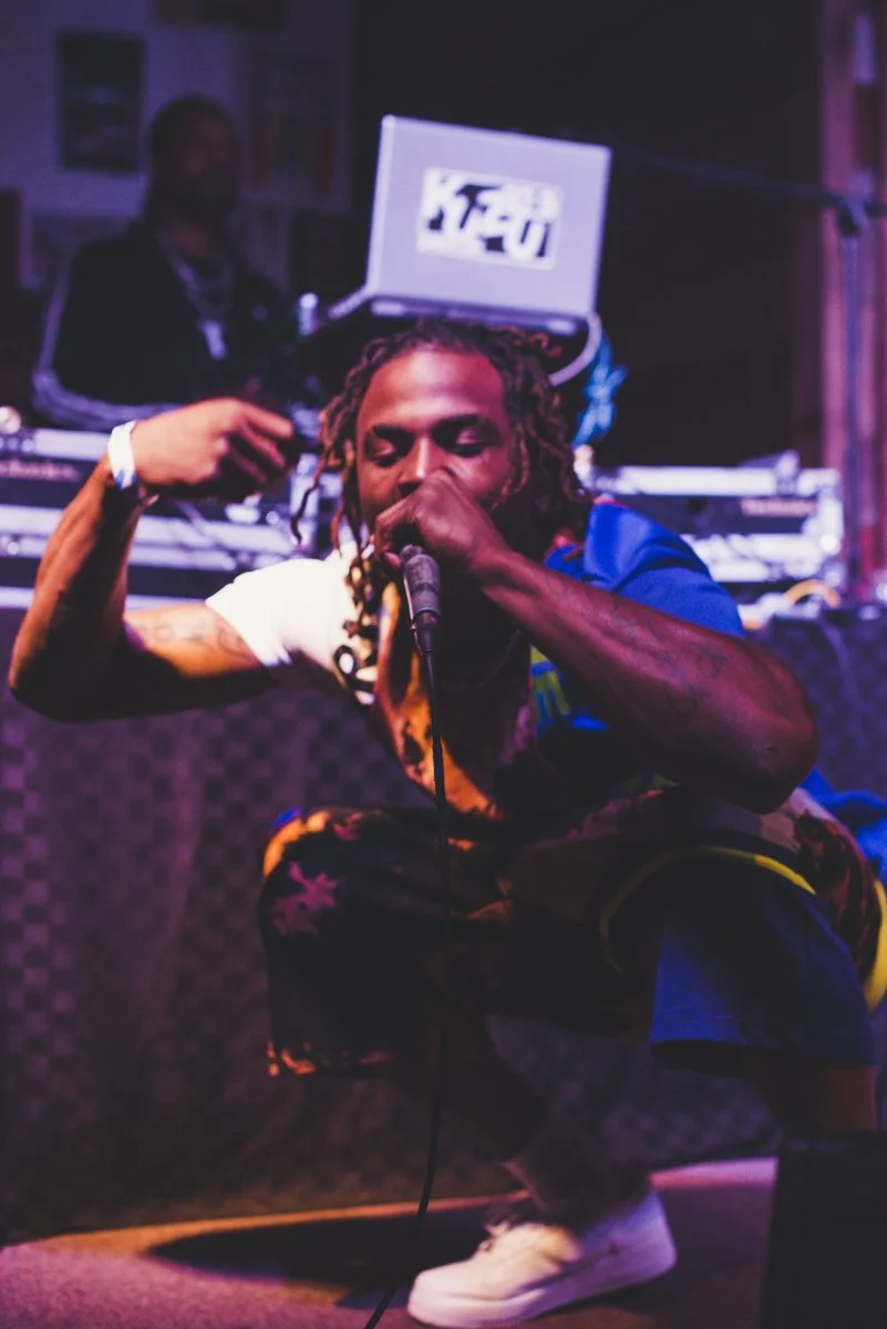 A rapper squatting on stage, holding a microphone close to his mouth, with a DJ in the background operating equipment and a laptop.