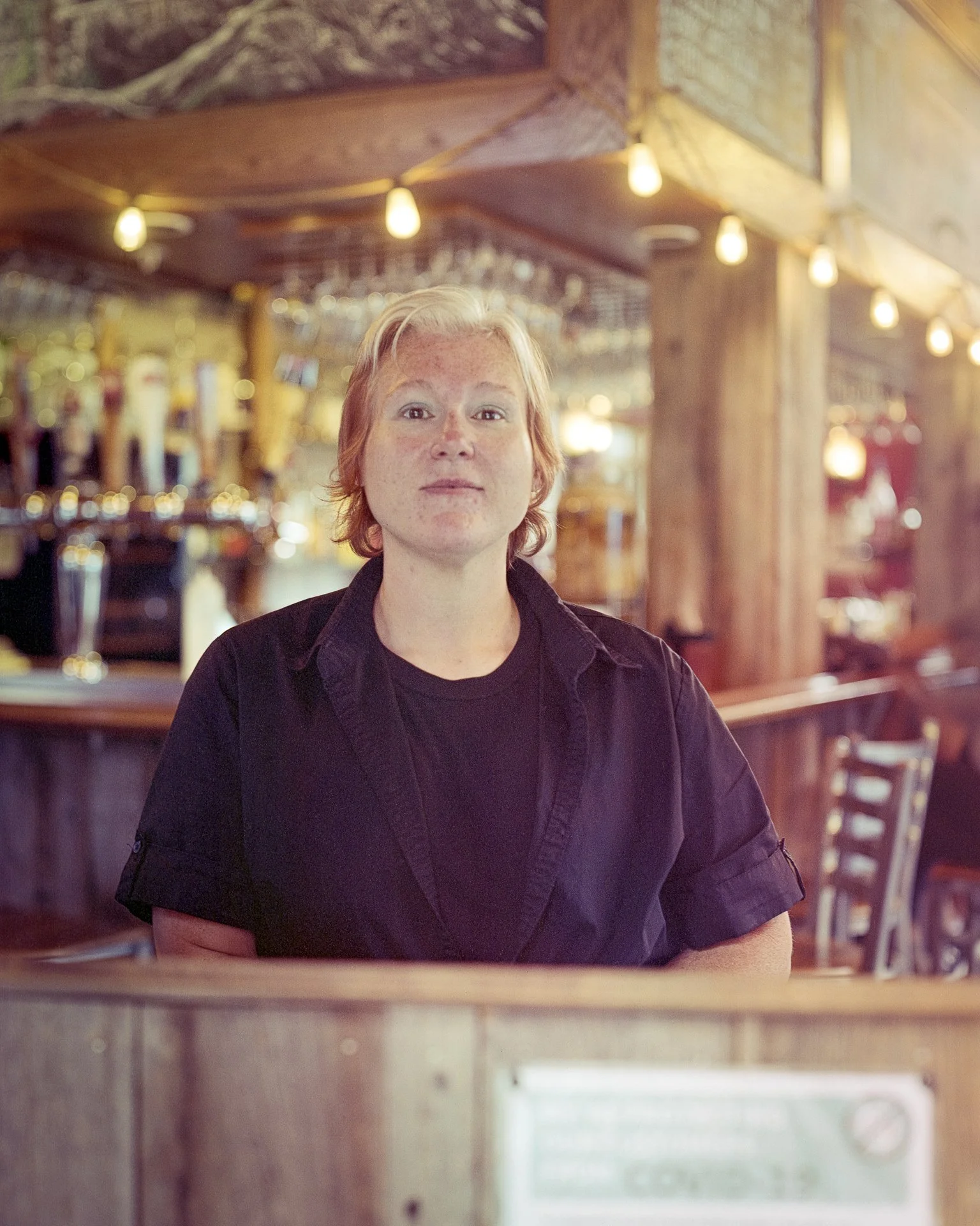 A woman with short, light-colored hair sitting in a rustic bar or restaurant with wooden interior and string lights.