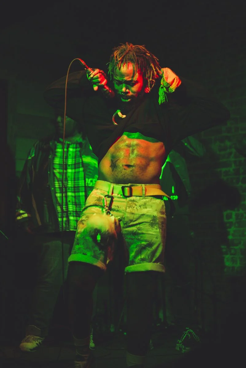 A performer with dreadlocks sings into a microphone with bright stage lighting, wearing shorts and a partially lifted shirt to reveal abs, while a person in plaid pants stands in the background.