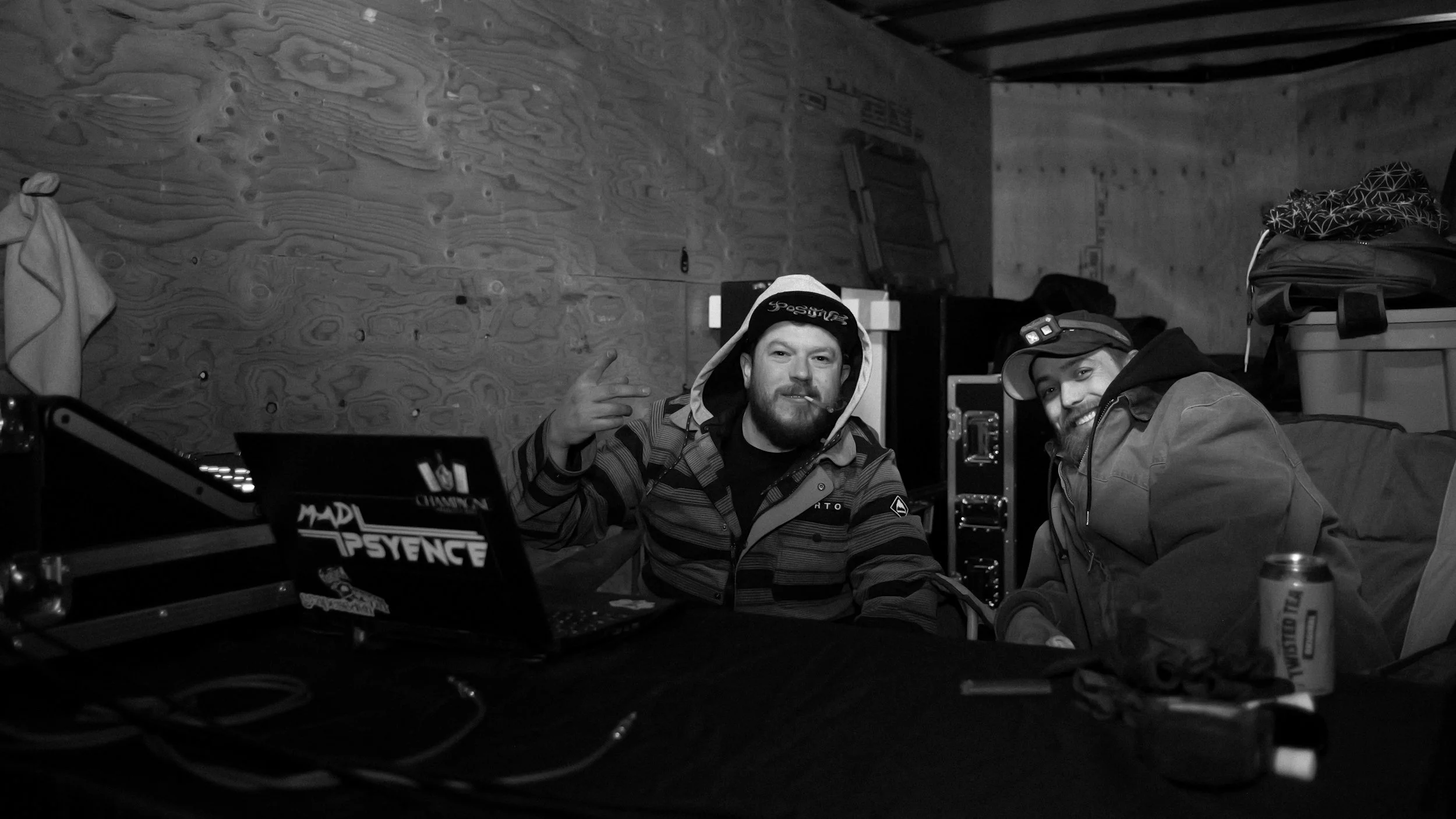 Two men sitting at a table in a wooden room, one making a peace sign and smiling, wearing a beanie and jacket, the other smiling and wearing a cap and jacket, surrounded by equipment and containers.