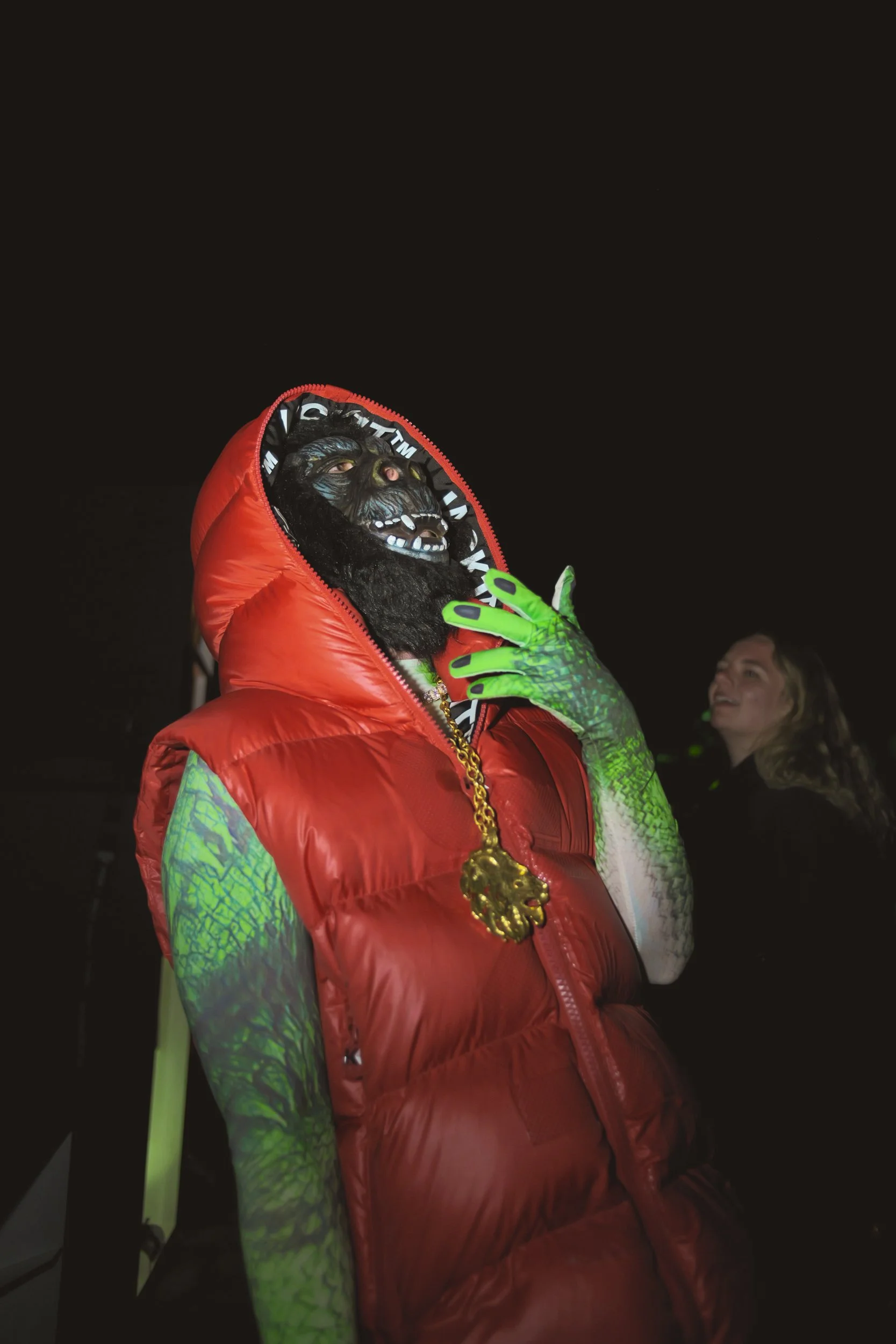 Person wearing a gorilla mask, red puffy vest, and neon green gloves, accessorized with a gold chain, at night with a woman smiling in the background.