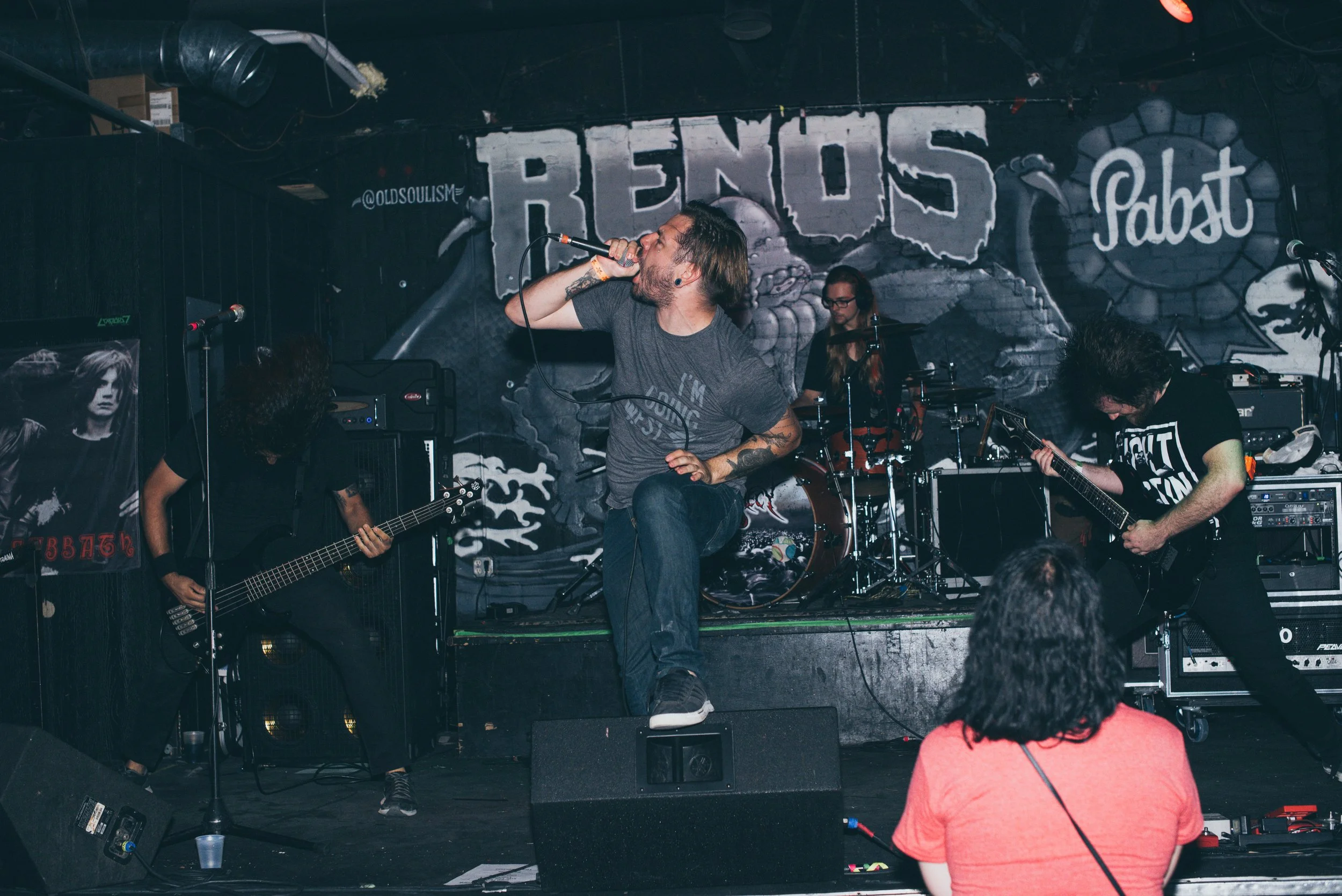 Band performing live on stage with a vocalist holding a microphone, guitarist, bassist, and drummer, with a person in the audience watching.