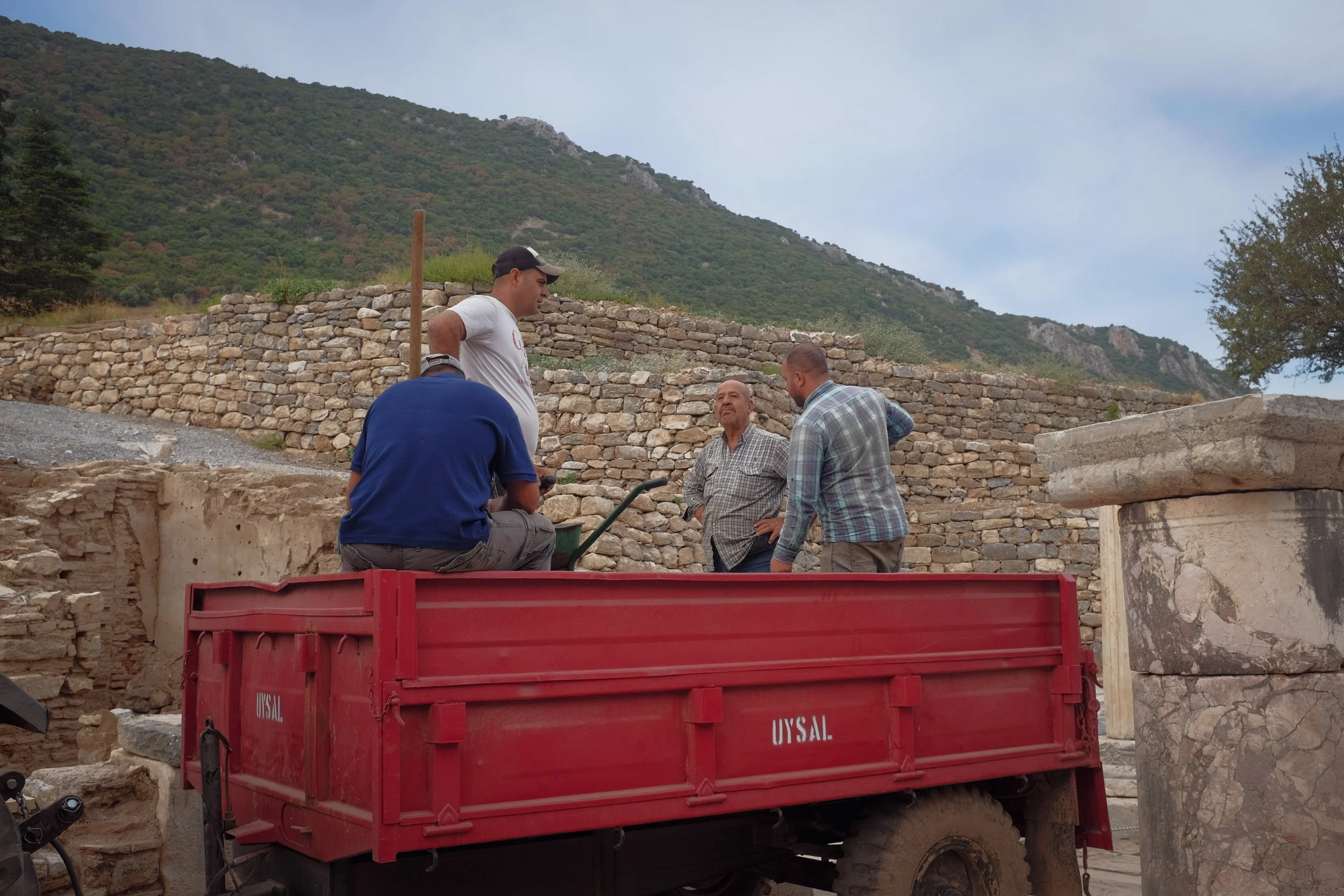 Four men discussing at an archaeological site with stone ruins and a hillside in the background, one sitting on a red truck.
