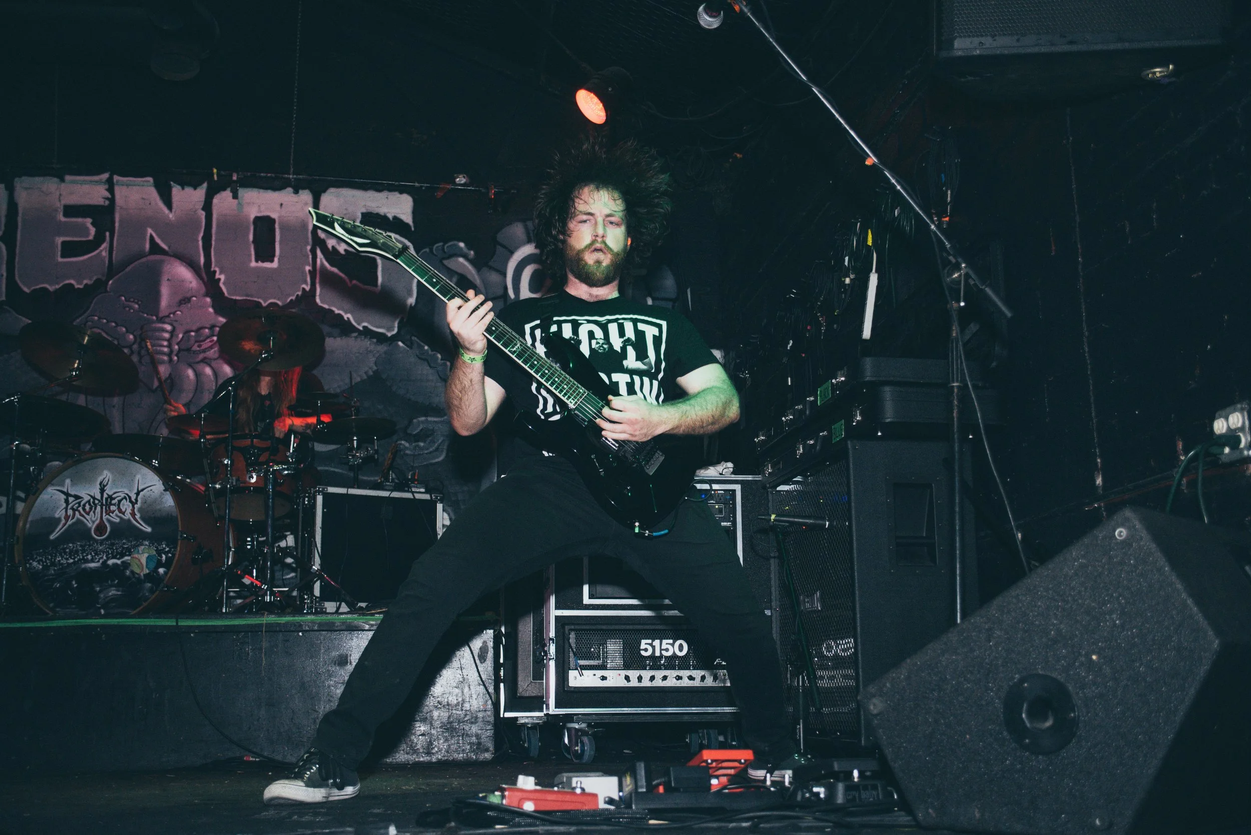A musician with curly hair and a beard playing an electric guitar on stage during a concert, with another drummer in the background, stage equipment, and a dark background.