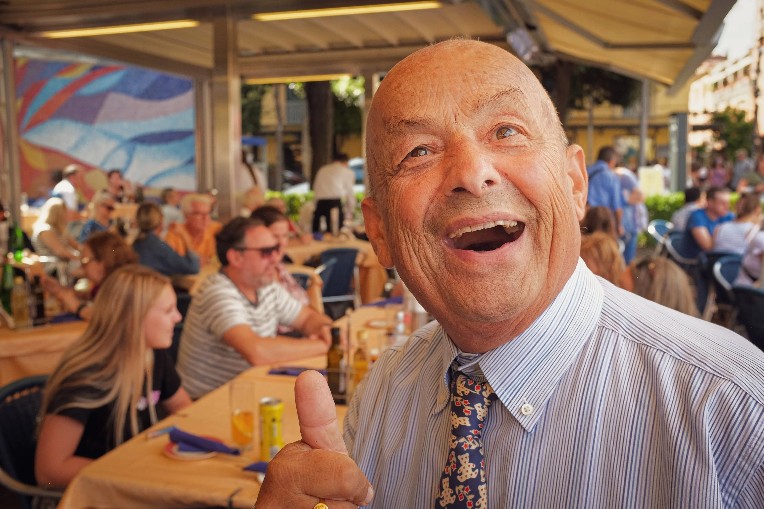 An elderly man with a bald head, smiling widely with his mouth open, giving a thumbs-up gesture at an outdoor social gathering or celebration with people seated at tables in the background.