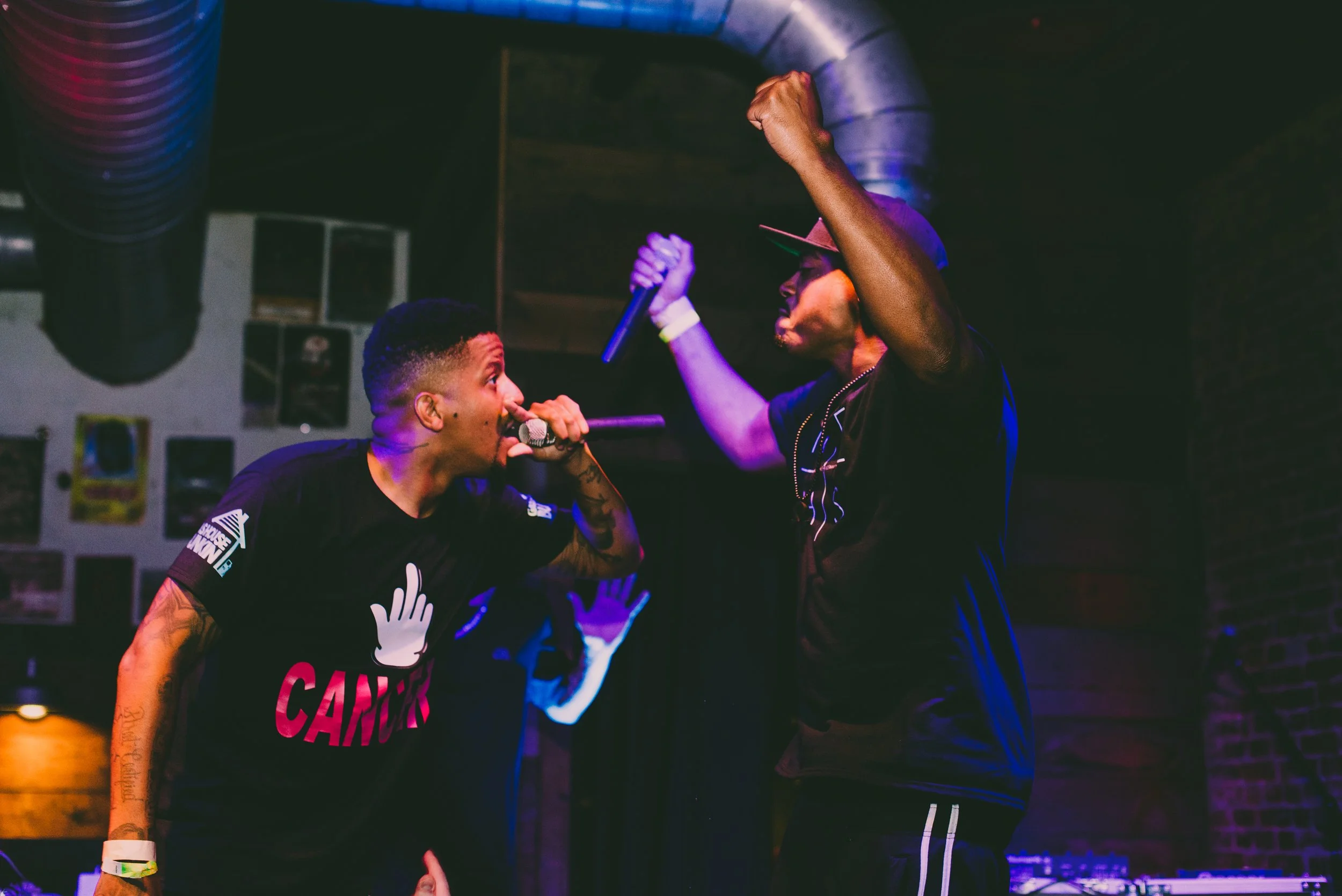 Two men performing on stage with microphones, one wearing a black shirt with a hand symbol and the word 'CANCER', and the other wearing a black shirt and cap, both raising their fists in energetic poses, in a dimly lit setting with colorful stage lig