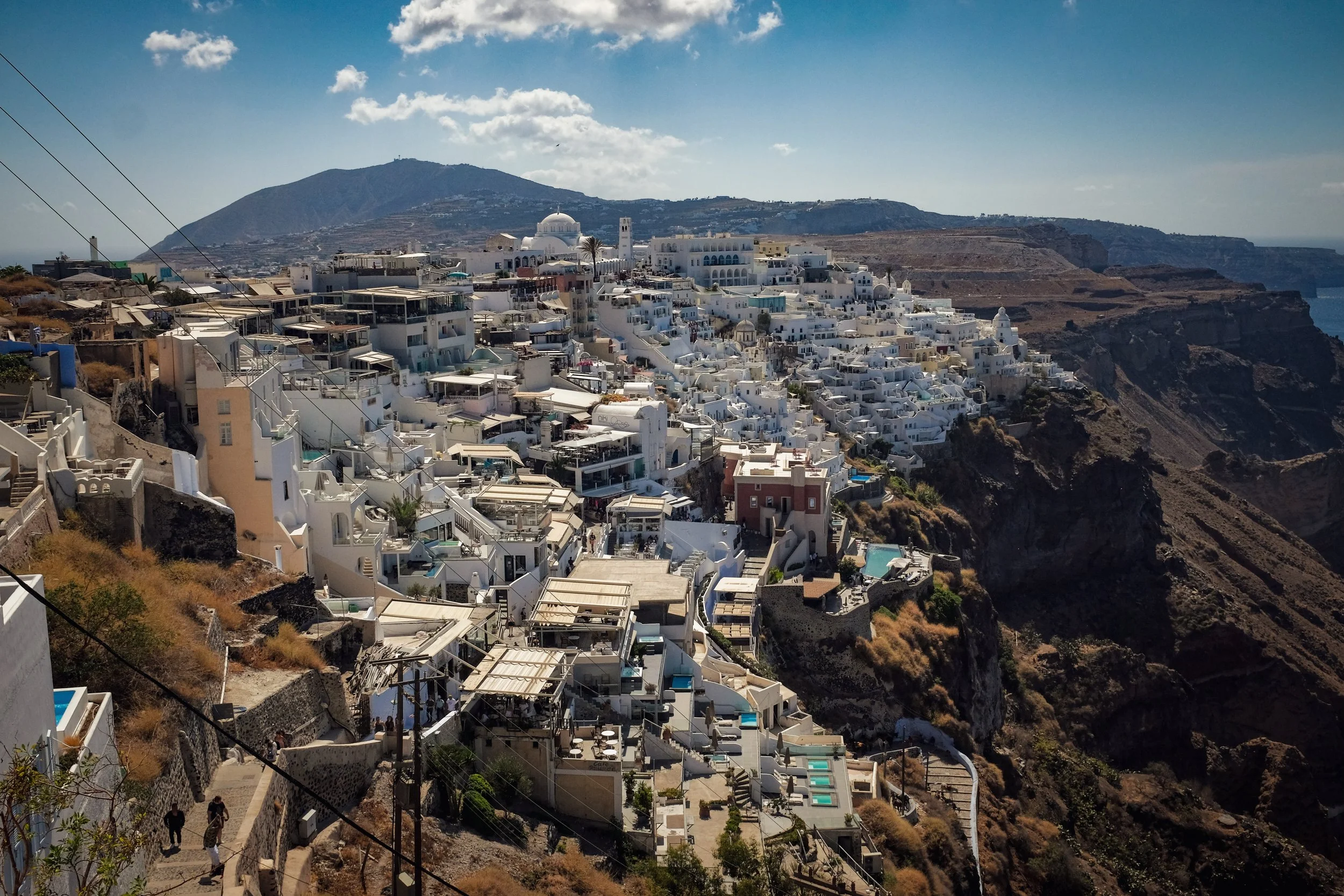 White buildings cascading down a cliffside on a hillside with a mountain in the background and a partly cloudy sky.