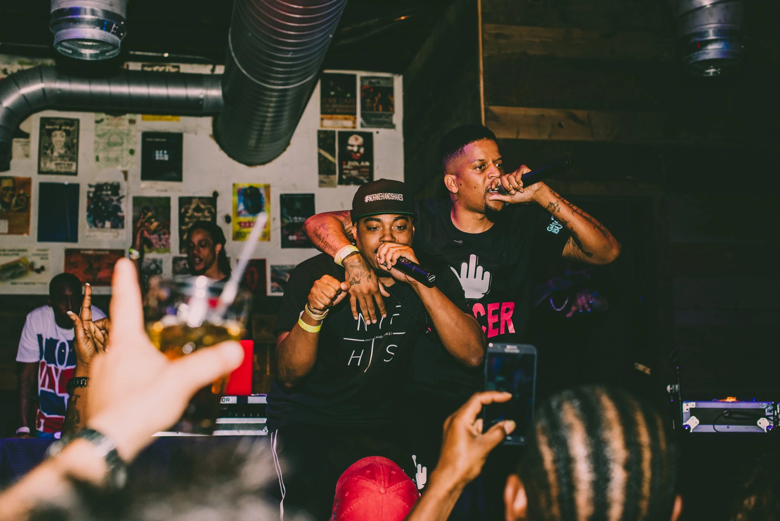 Two male performers singing into microphones on stage, one with arm around the other, at a concert or club. Several audience members are visible in the foreground, one taking a photo with a smartphone, and one holding a drink. The background features