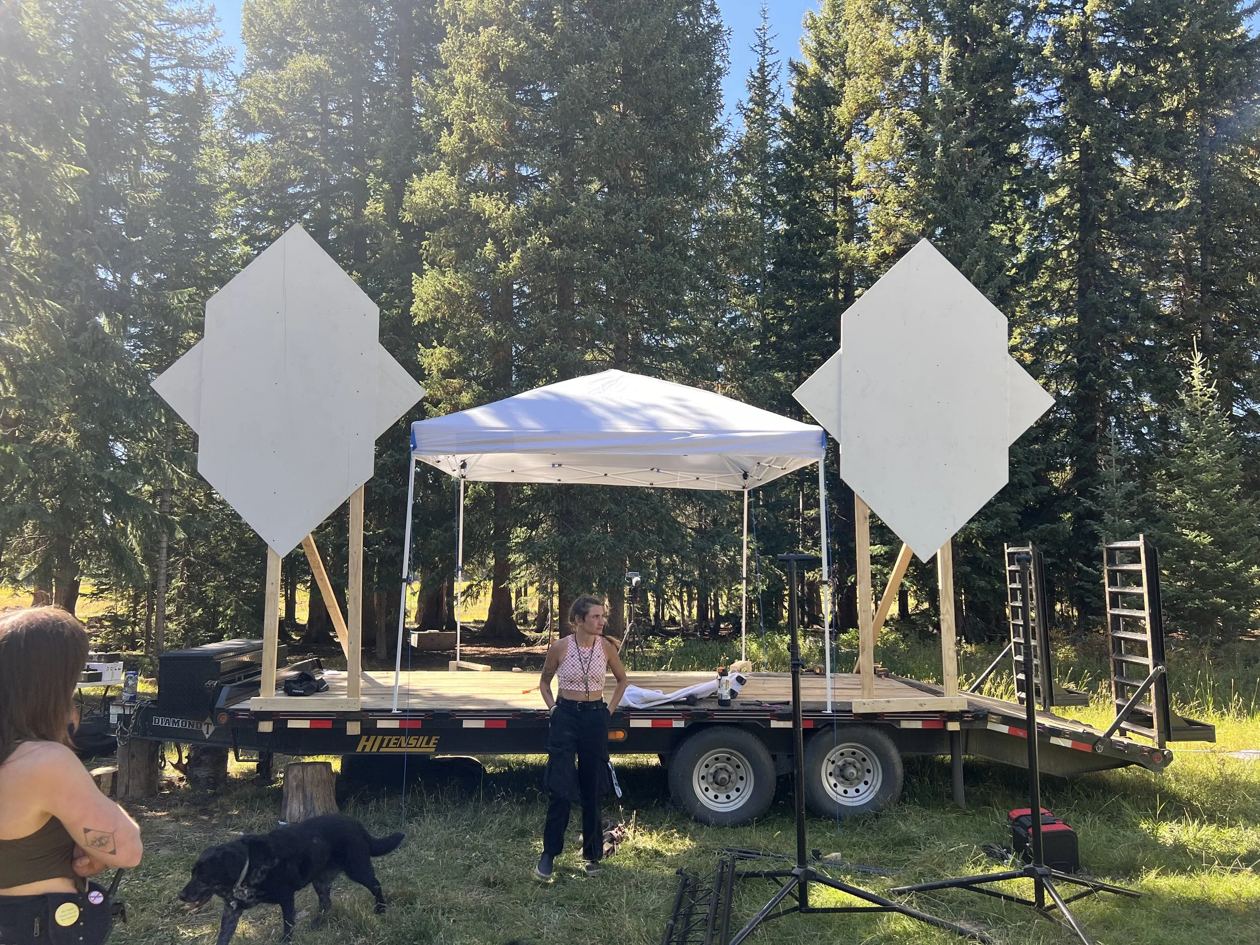 A woman stands on a trailer with two large white directional signs and a white canopy tent, surrounded by trees in a forest setting. Another woman with a tattoo and a black dog are nearby.
